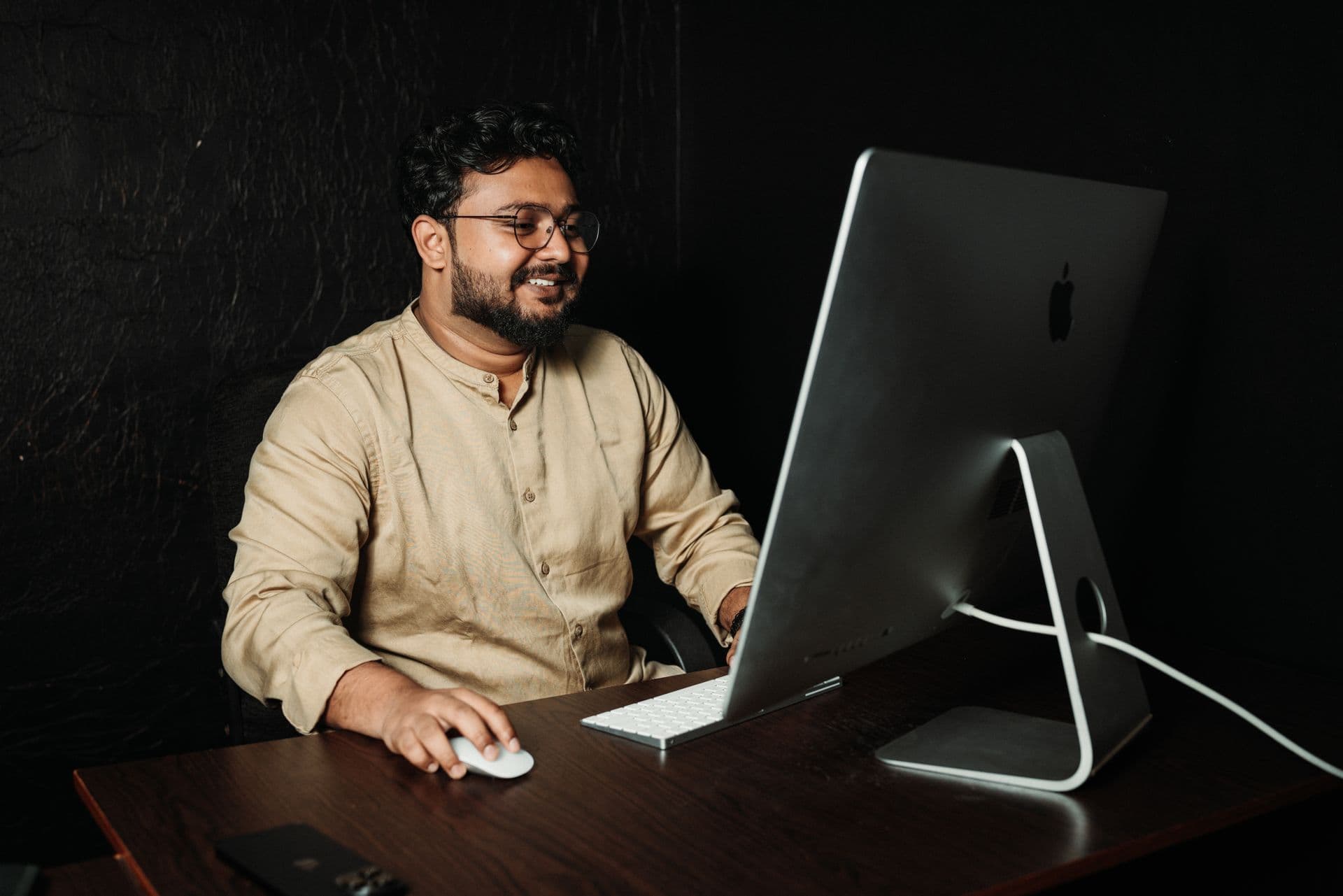 Smiling man in a beige band-collar shirt using a desktop computer in a dark room