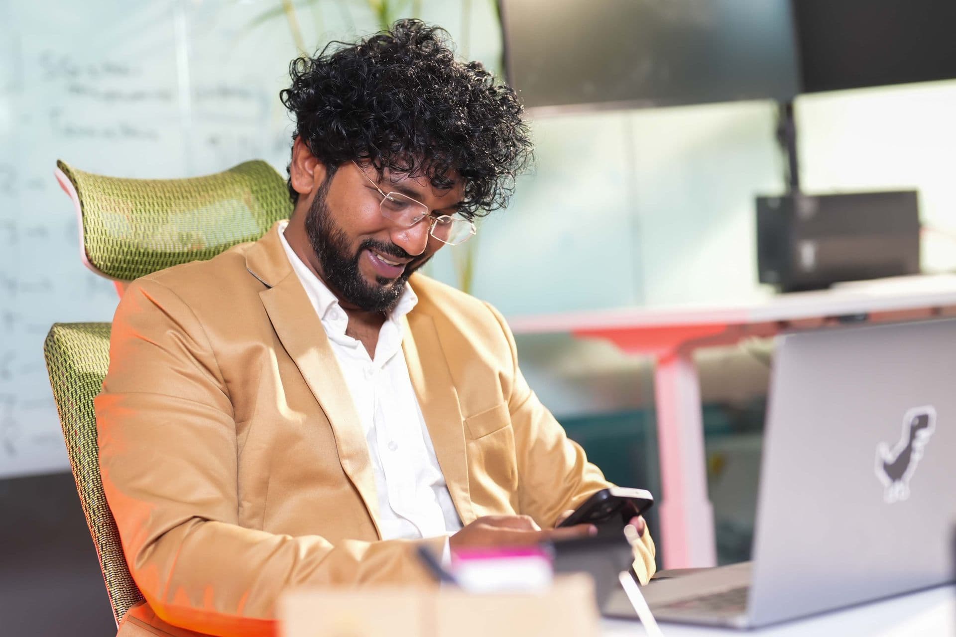 Man in a tan blazer smiles at his smartphone beside an open laptop