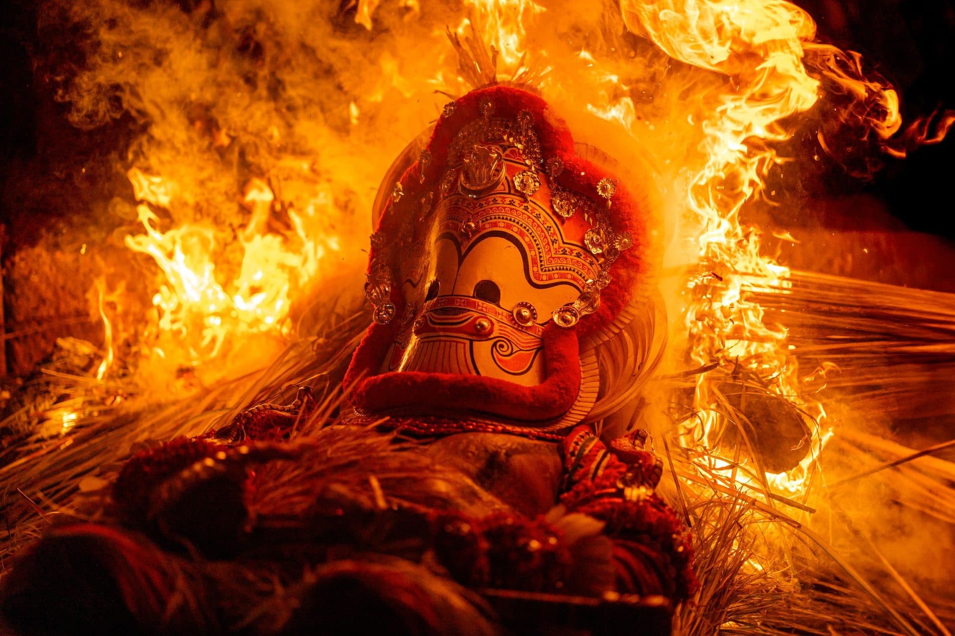 Theyyam performer's ornate red headdress and painted face amid roaring flames at night