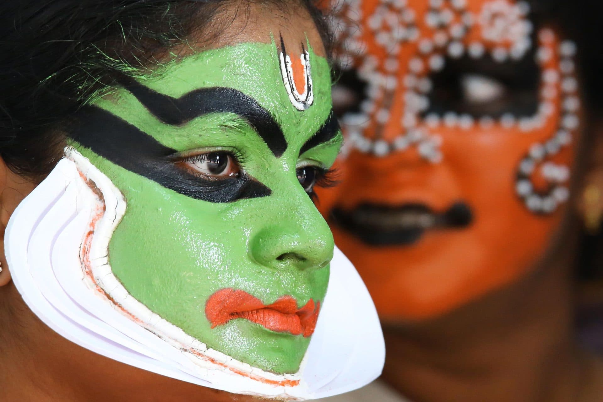 Green Kathakali face paint and white chutti in close-up, a second painted face behind