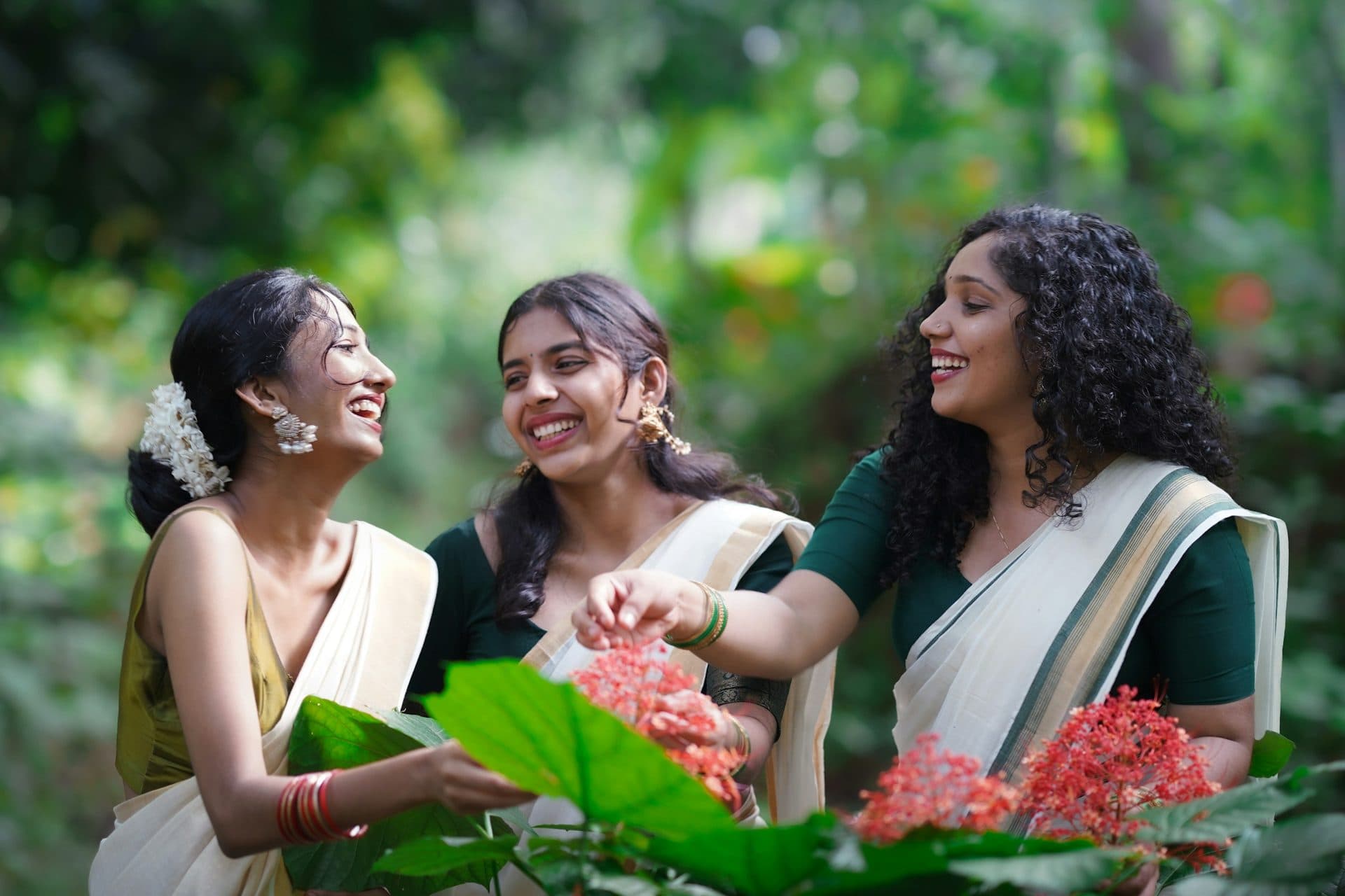 Three women in cream sarees with gold and green borders laugh beside red flowers