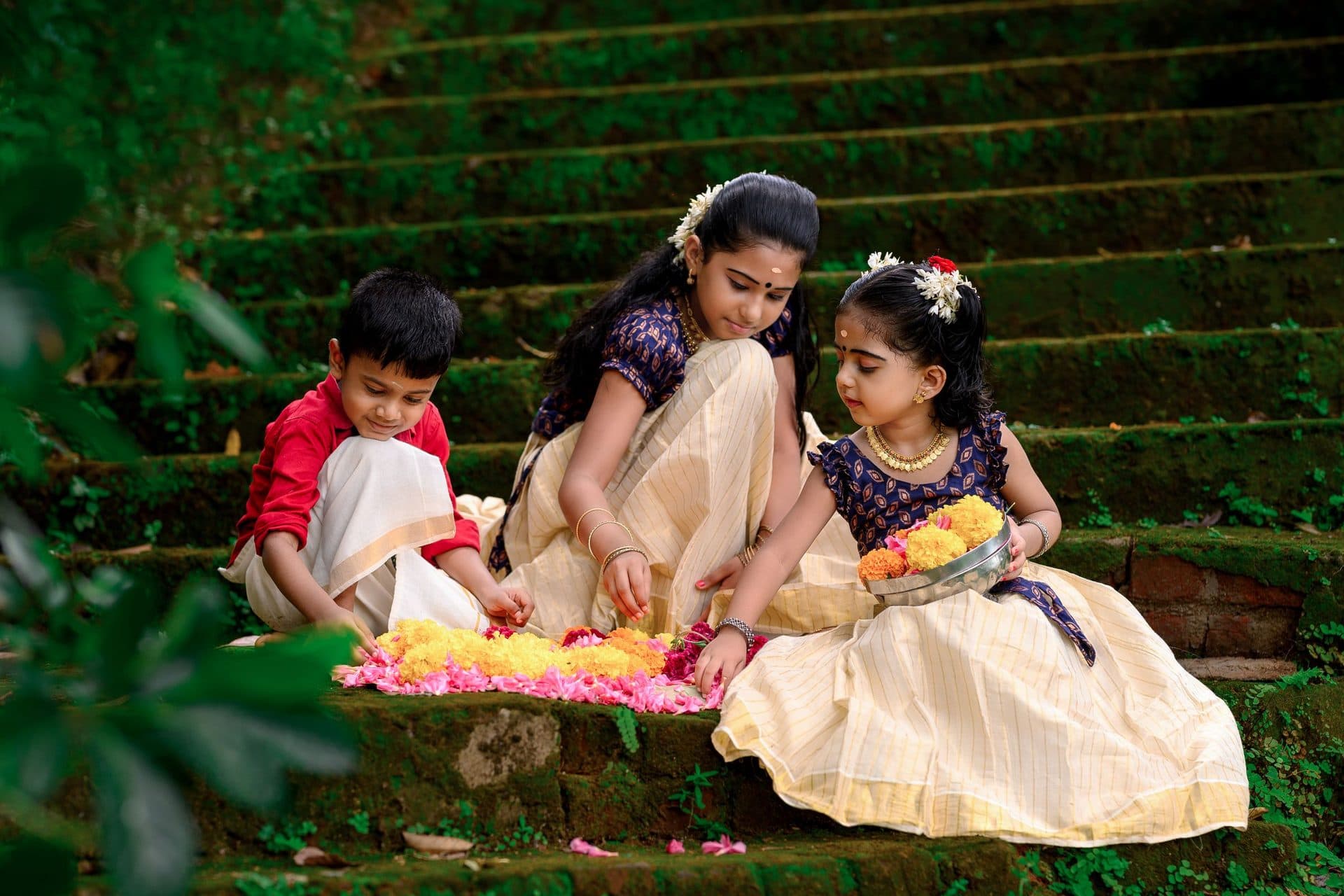 Three children in cream festive clothes arrange a flower carpet on mossy stone steps