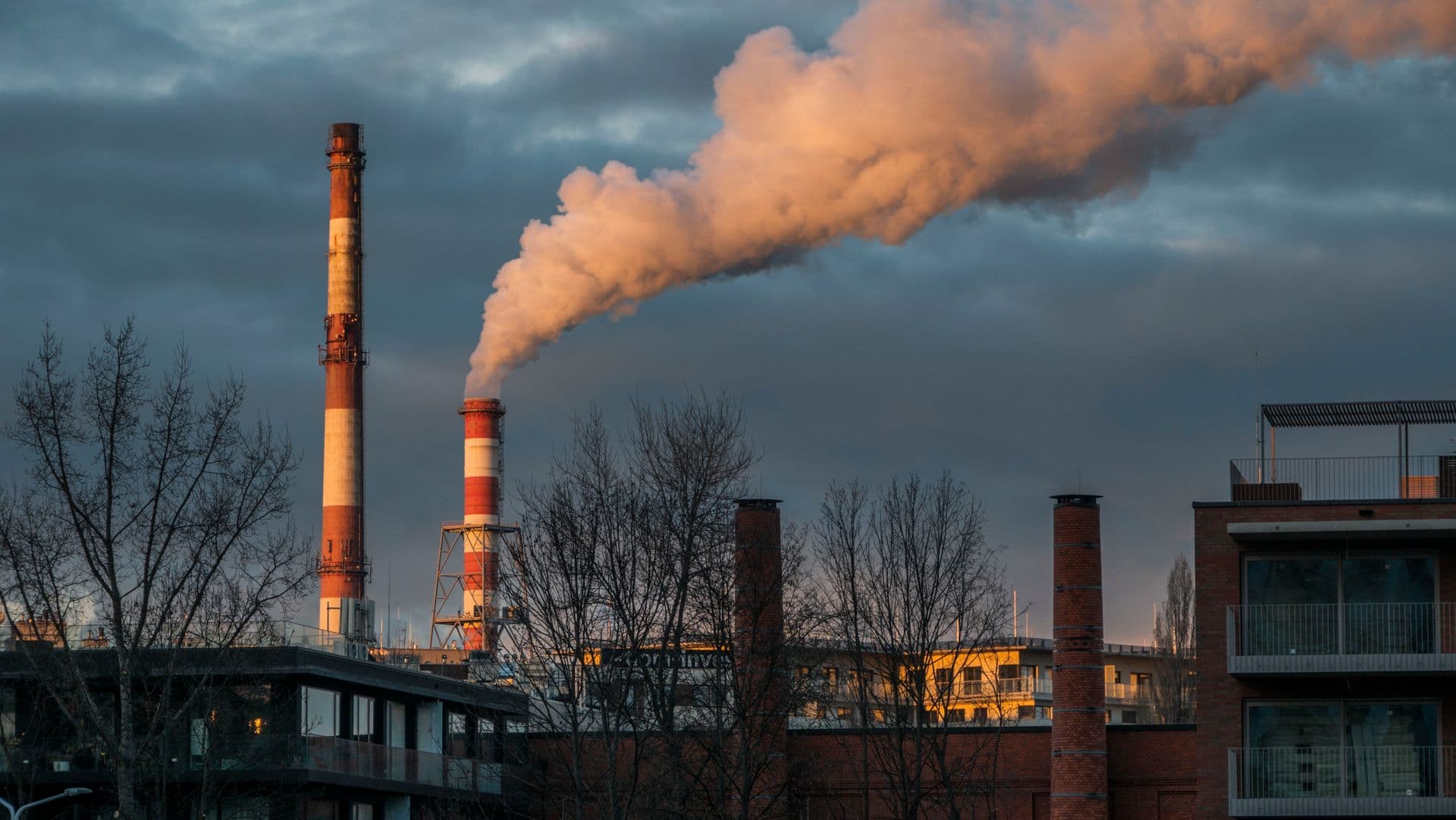 Industrial chimneys releasing smoke into a cloudy sky above buildings and bare trees