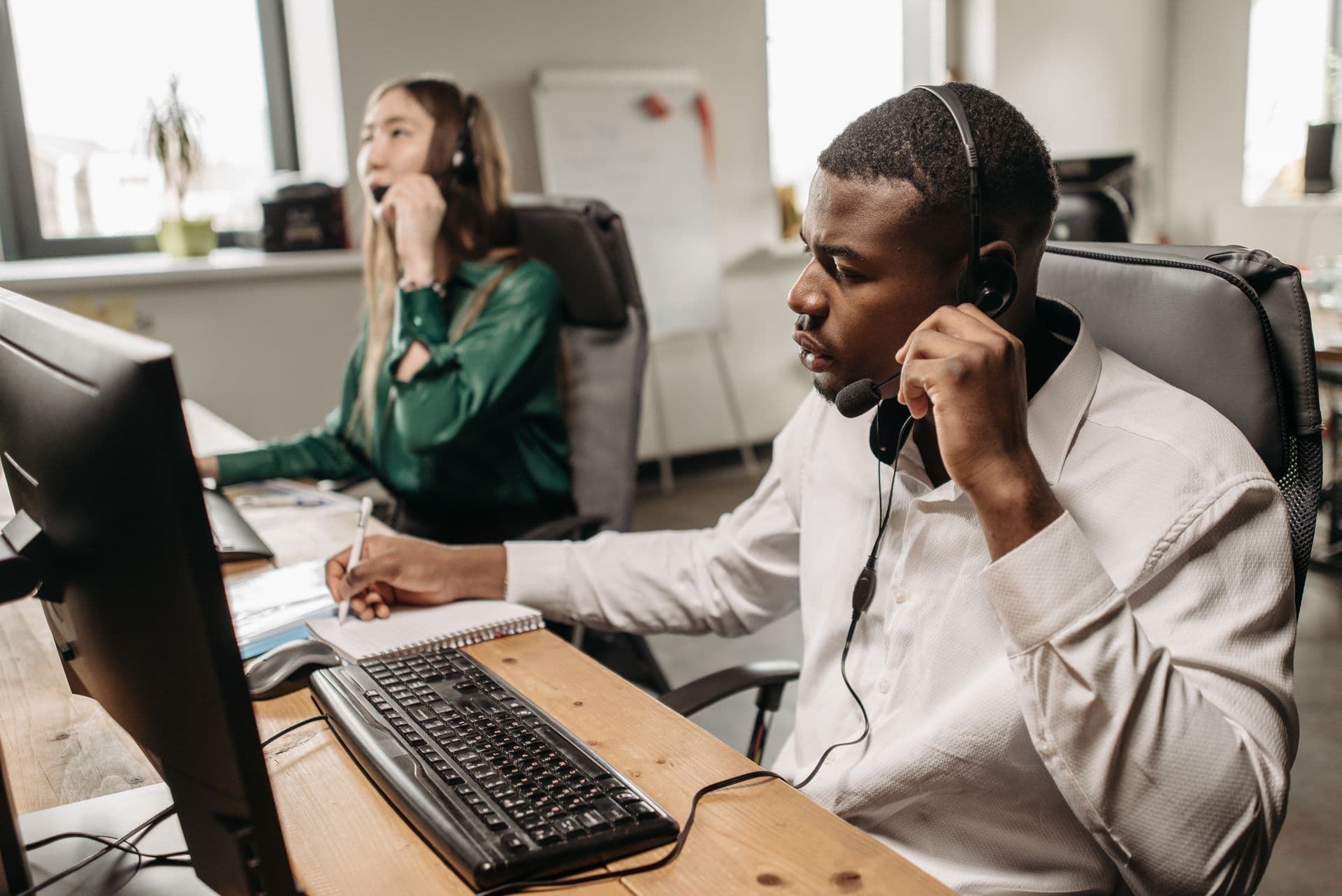 Two colleagues on headset calls at computers, one taking notes, in a bright office