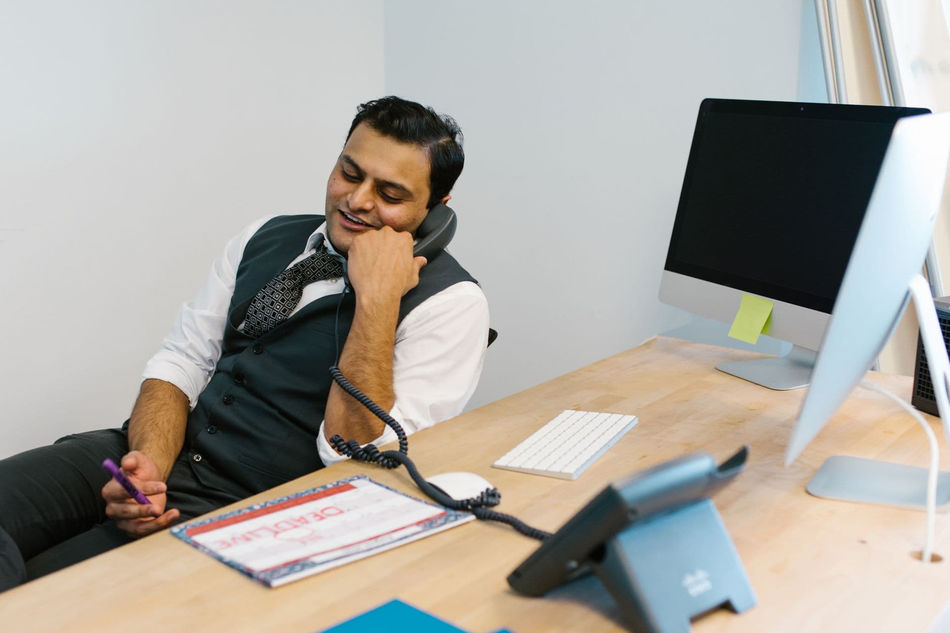 Smiling man in a waistcoat talks on a desk phone while taking notes