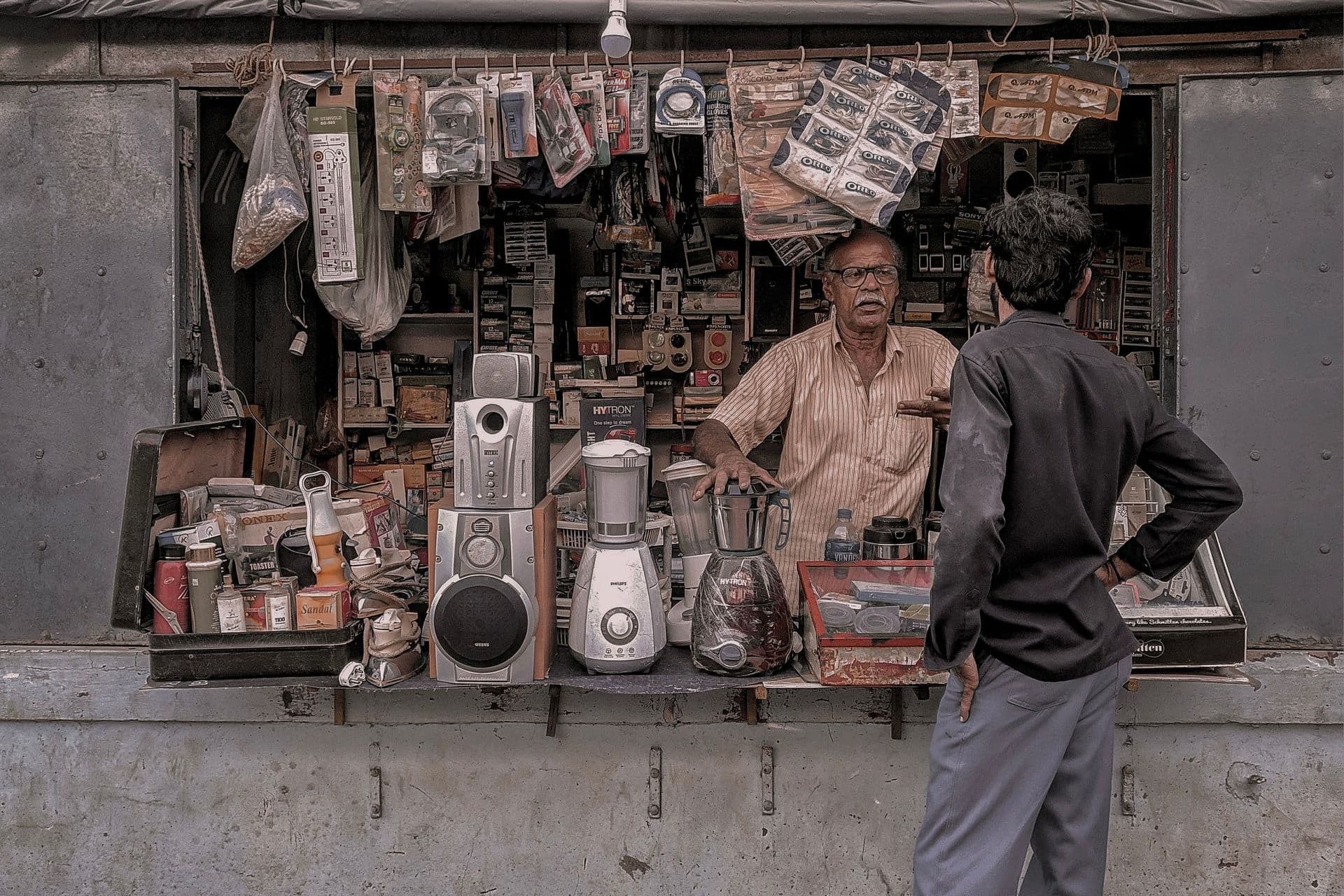 Shopkeeper talks with a customer at a small stall packed with speakers, mixers and gadgets