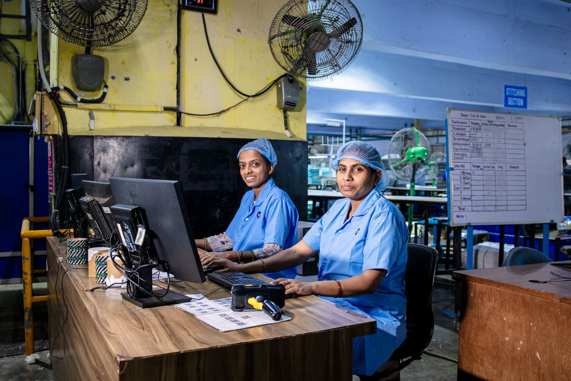 Two women in blue uniforms and hairnets working at computers in a factory office