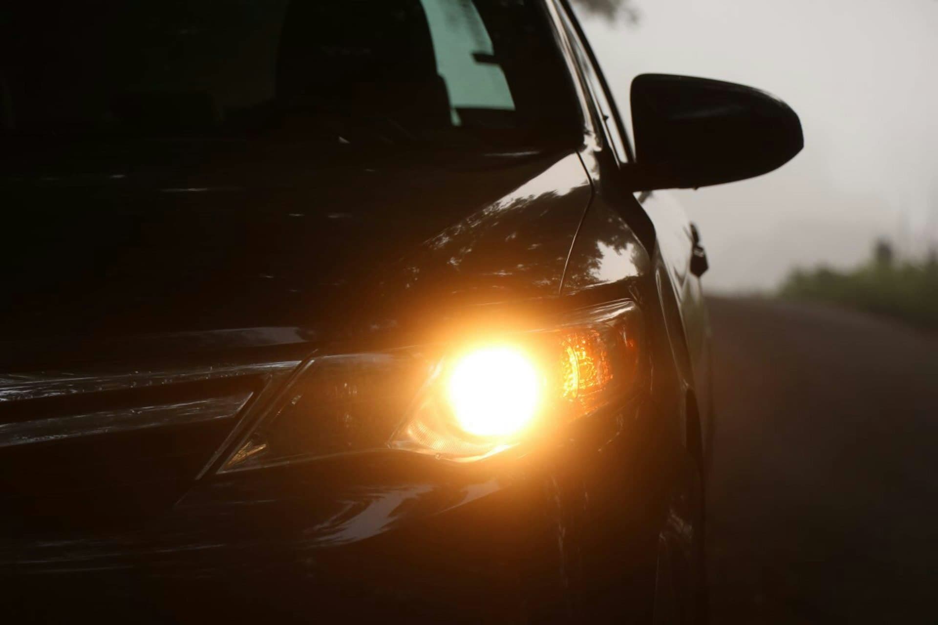 Close-up of a dark car's glowing amber headlight on a foggy road at dusk