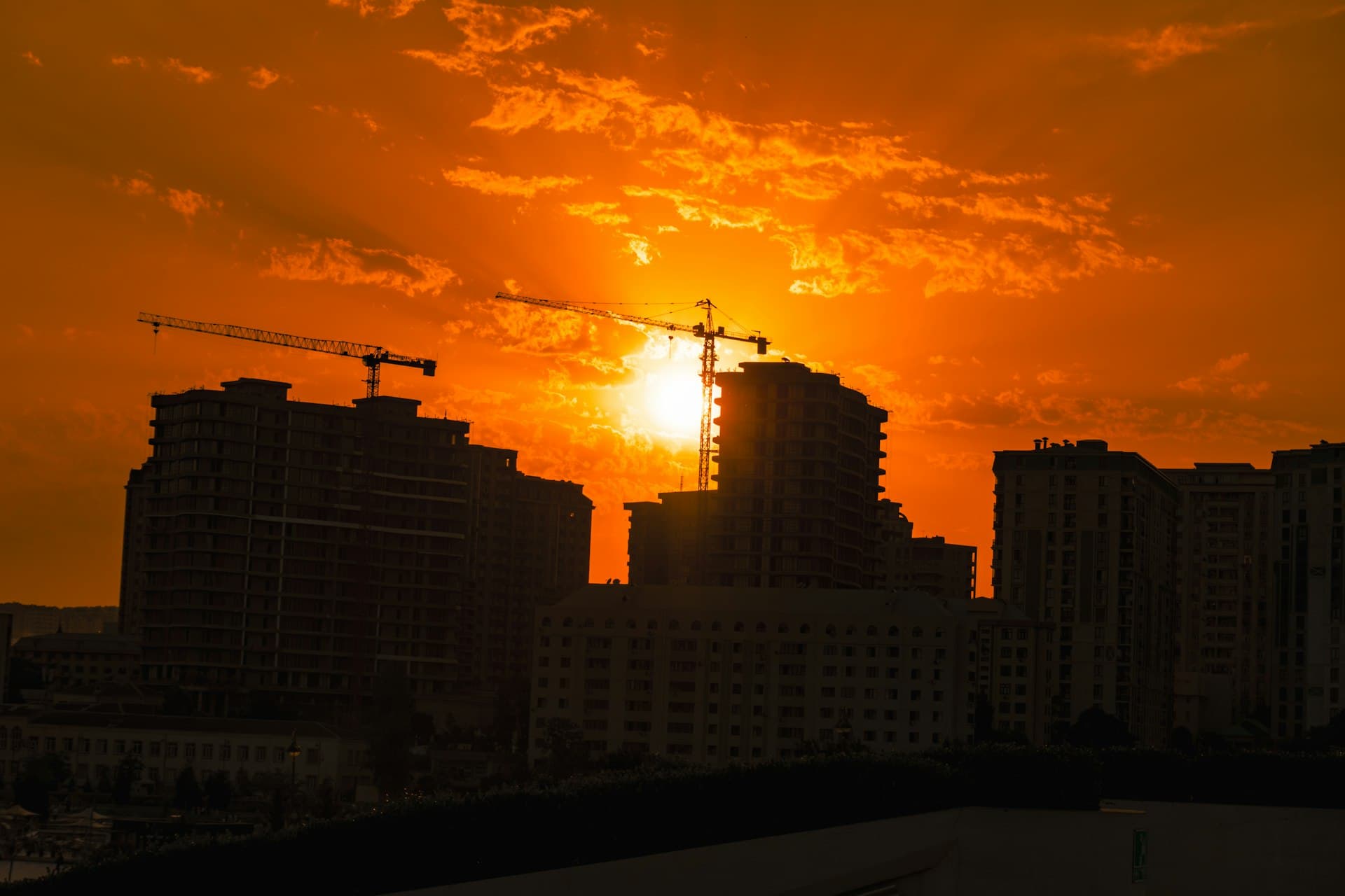 Construction cranes and unfinished high-rise buildings silhouetted against a fiery orange sunset sky