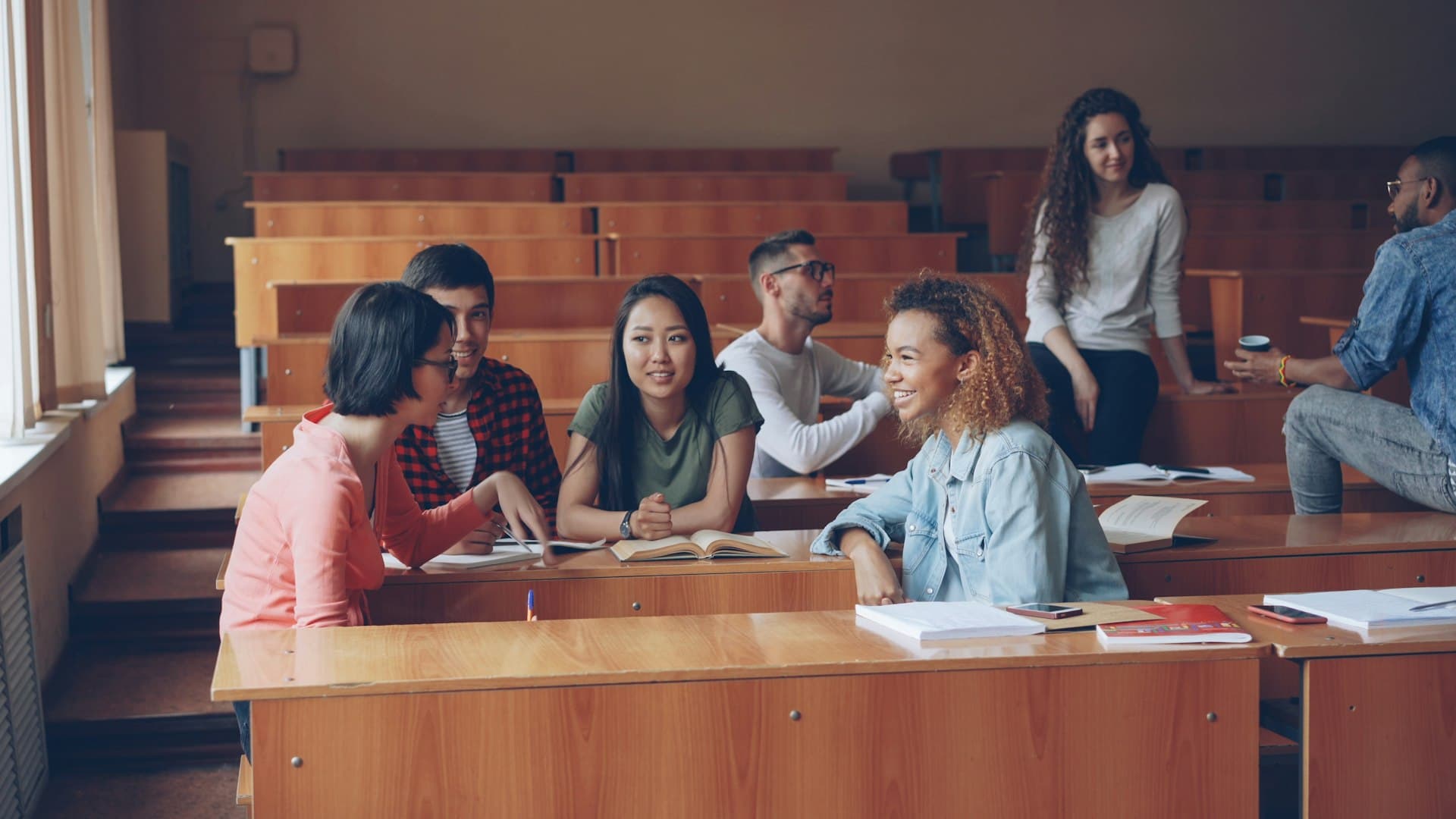 University students chatting and studying together at wooden desks in a tiered lecture hall