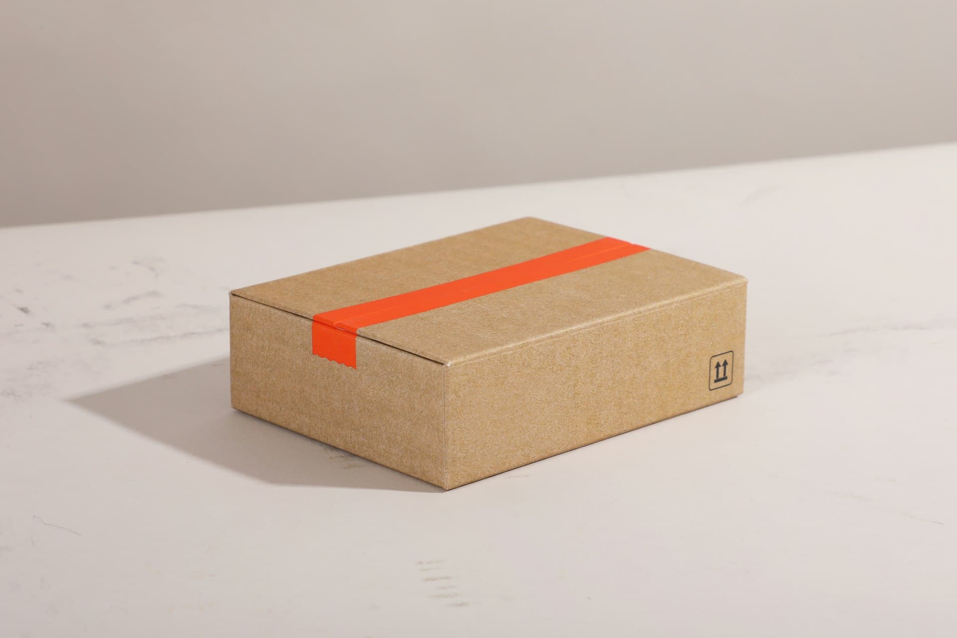 Brown cardboard shipping box sealed with bright orange tape on a light marble surface