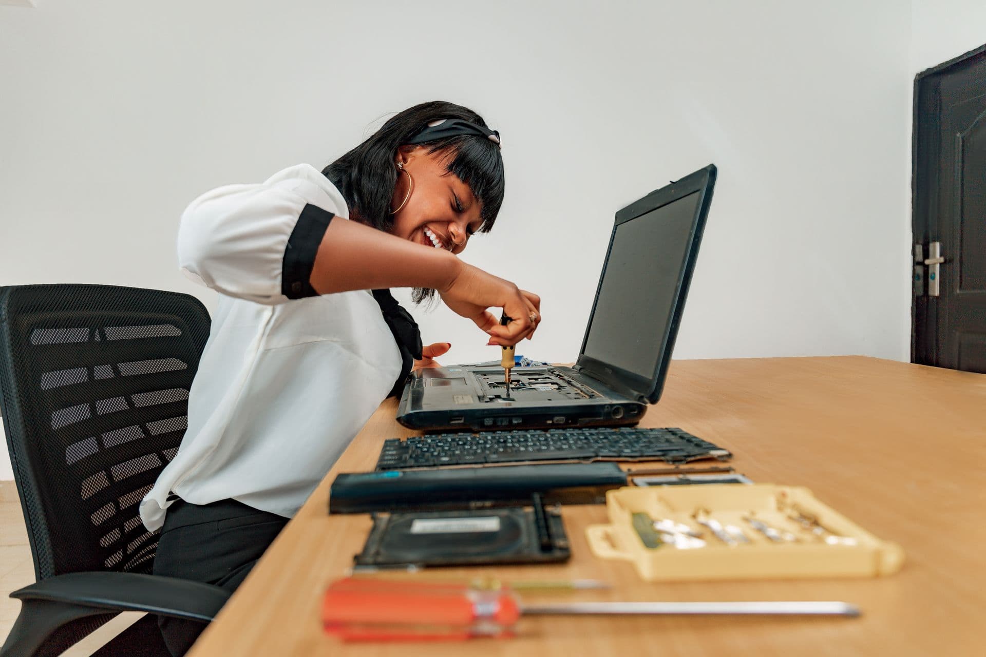 Woman working on a laptop chassis with a screwdriver beside removed keyboard and parts
