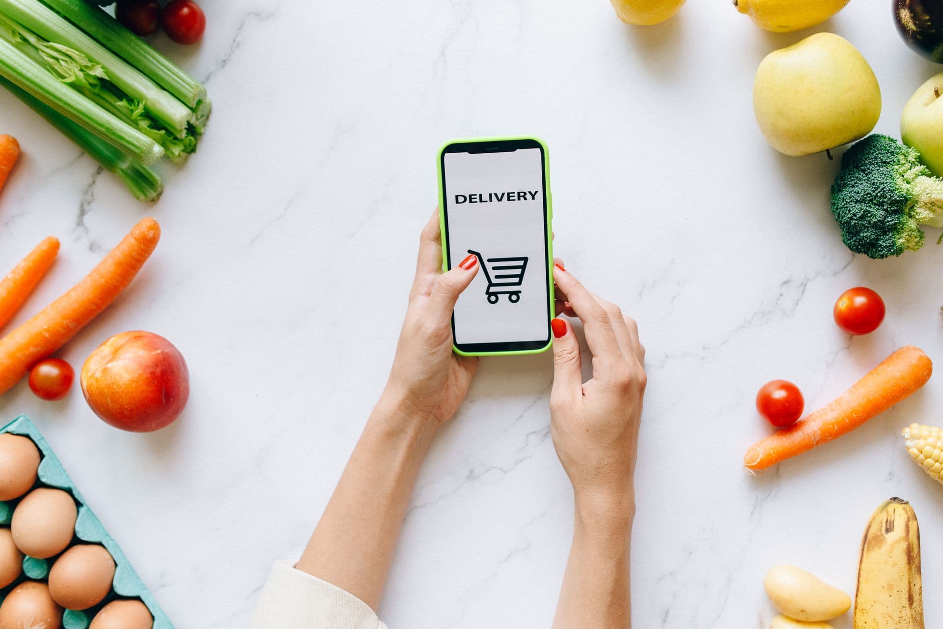 Hands holding a smartphone showing a grocery delivery app among fresh fruit and vegetables