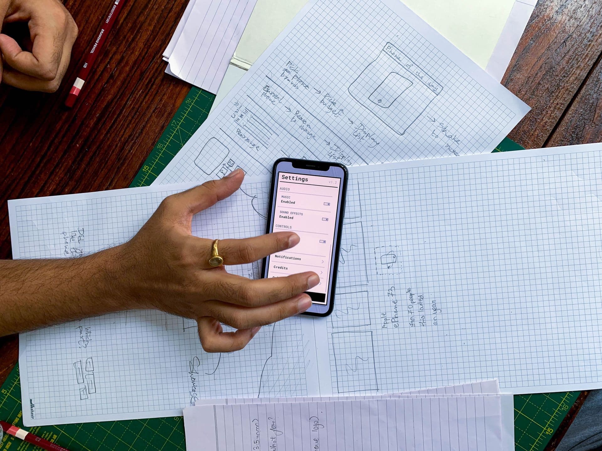 Hand holding a smartphone with a settings screen over hand-drawn app wireframes
