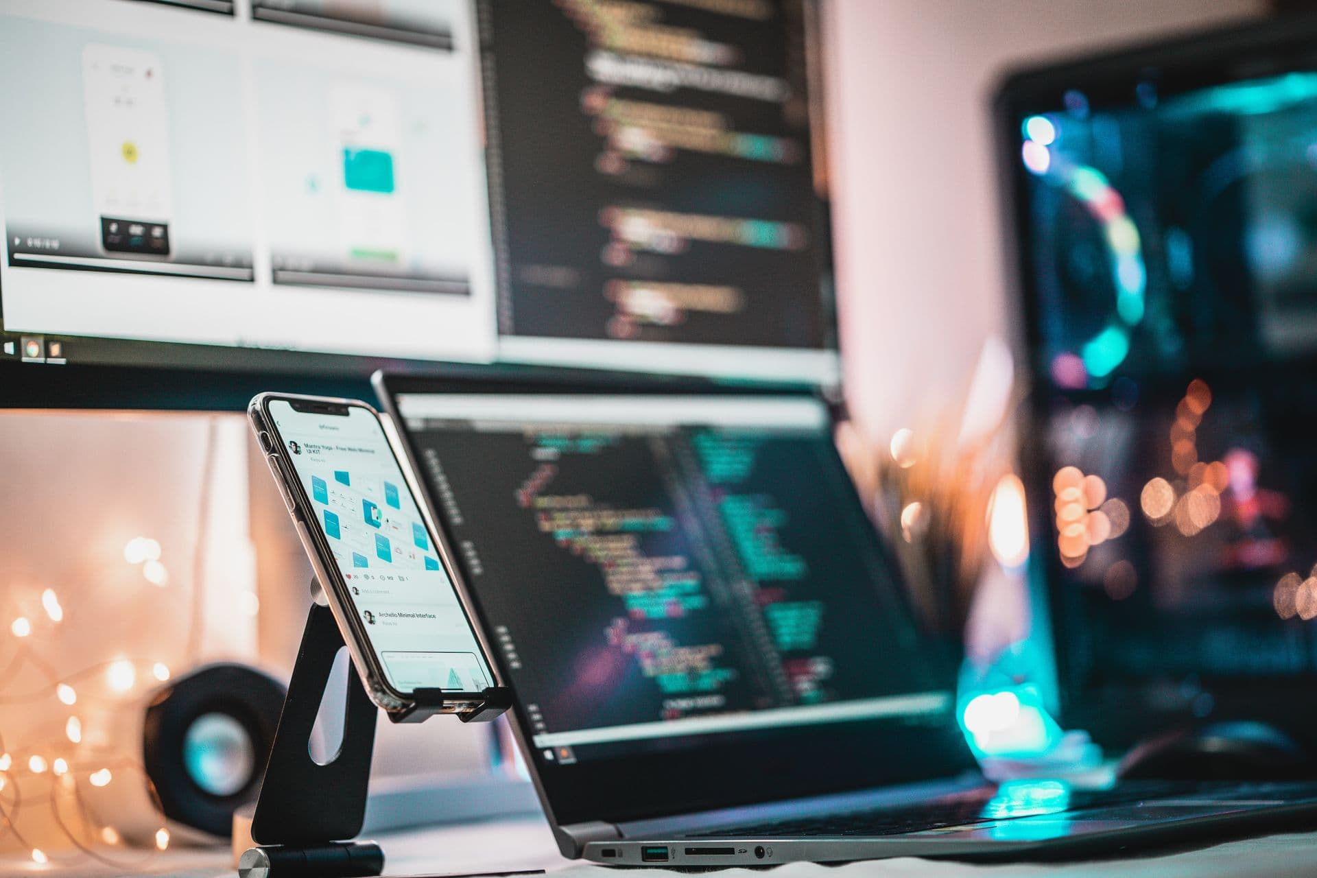 Smartphone on a desk stand beside a laptop and monitor displaying code