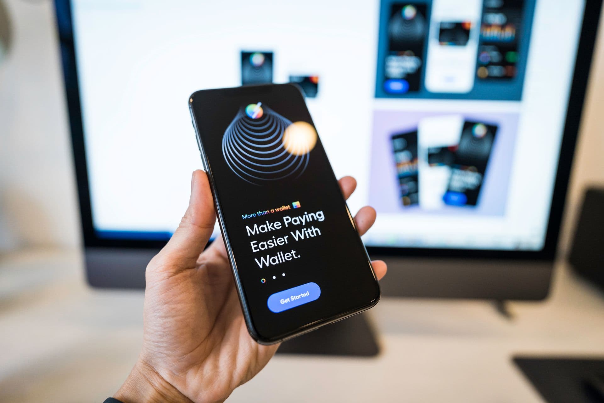 Hand holding a phone showing a wallet app, with app designs on a monitor behind