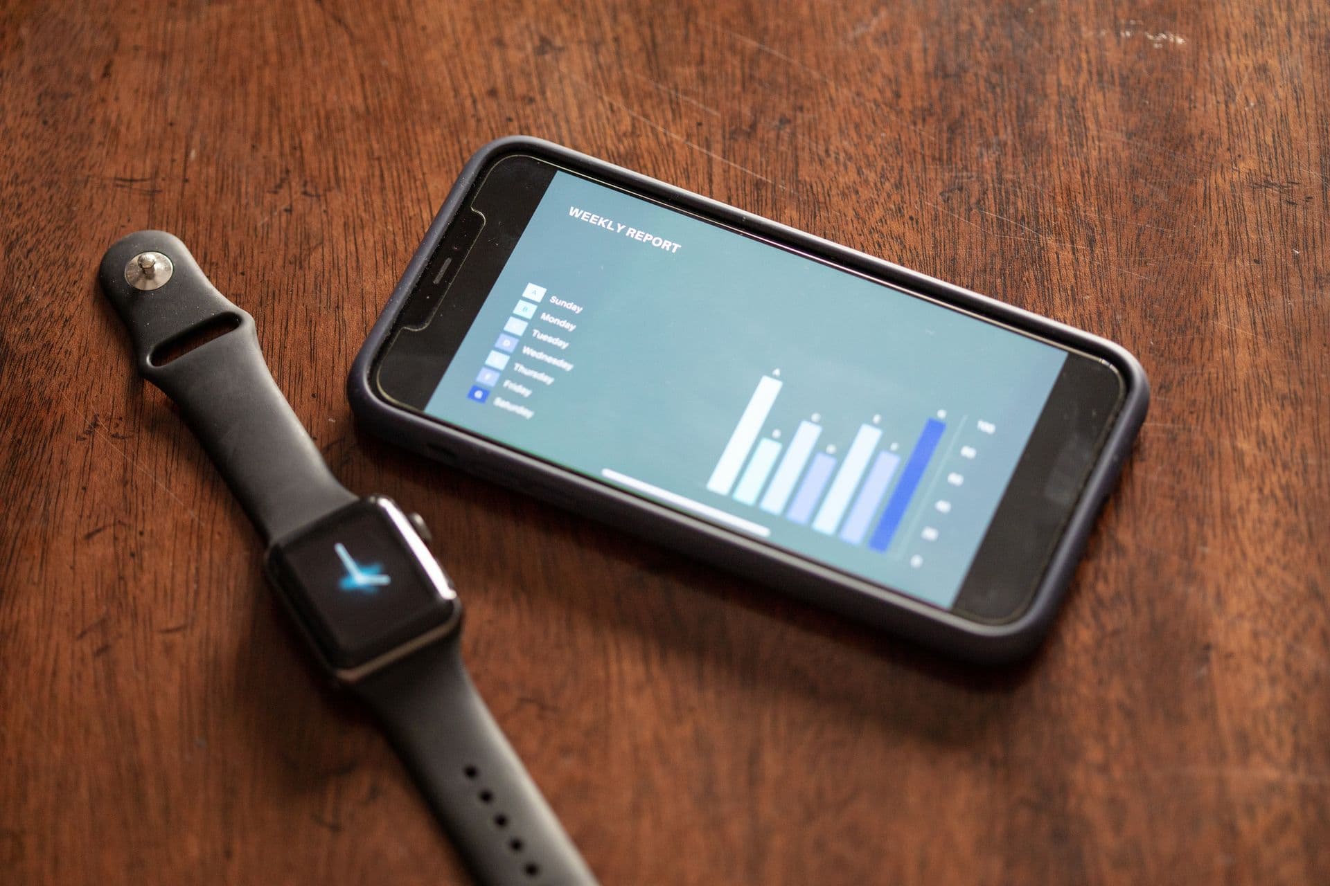 Smartwatch beside a smartphone showing a weekly report bar chart on a wooden table