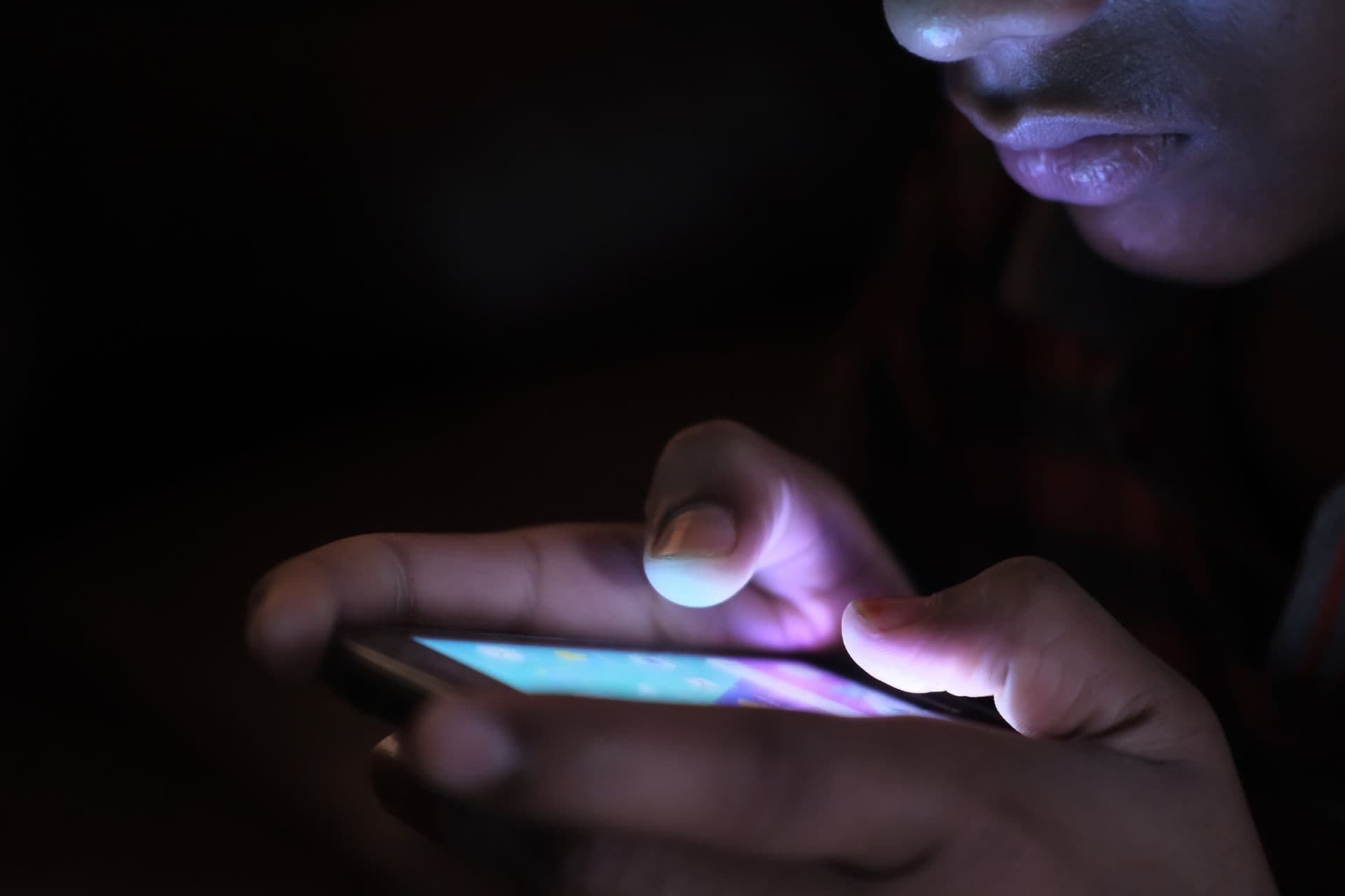 Person tapping a glowing smartphone screen in a dark room, hands lit by the display