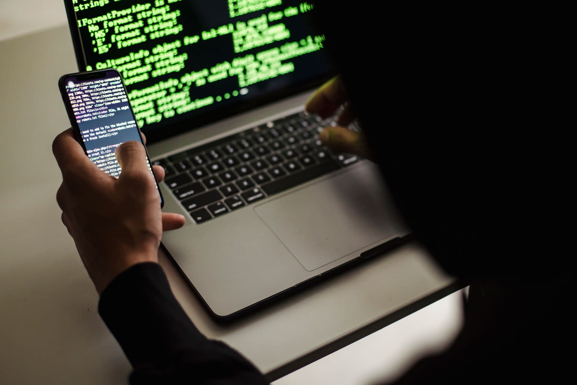 Developer holding a smartphone showing code while a laptop displays green code in a dark room