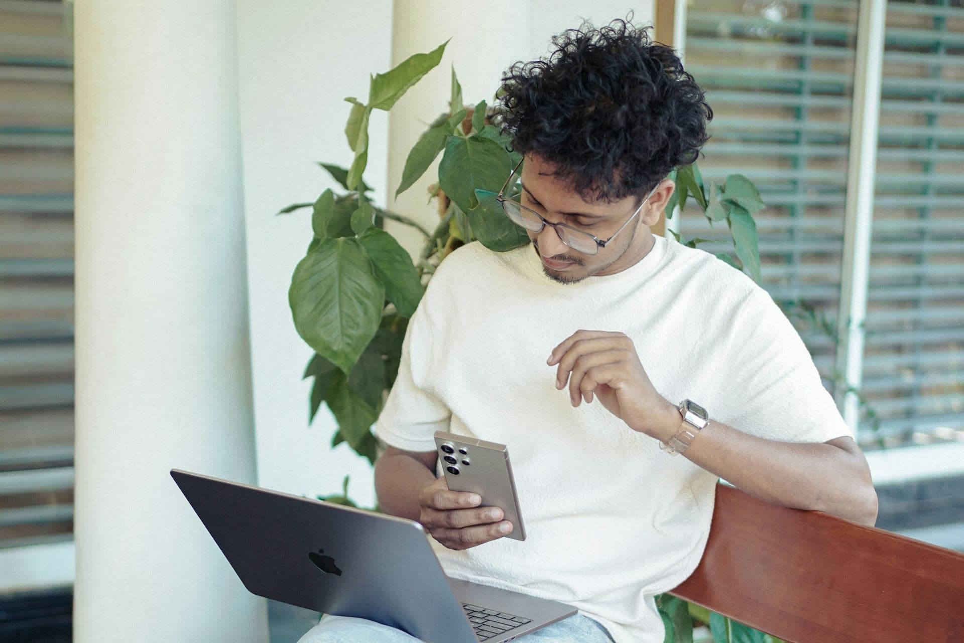 Young man in glasses checks his smartphone with a laptop on his lap