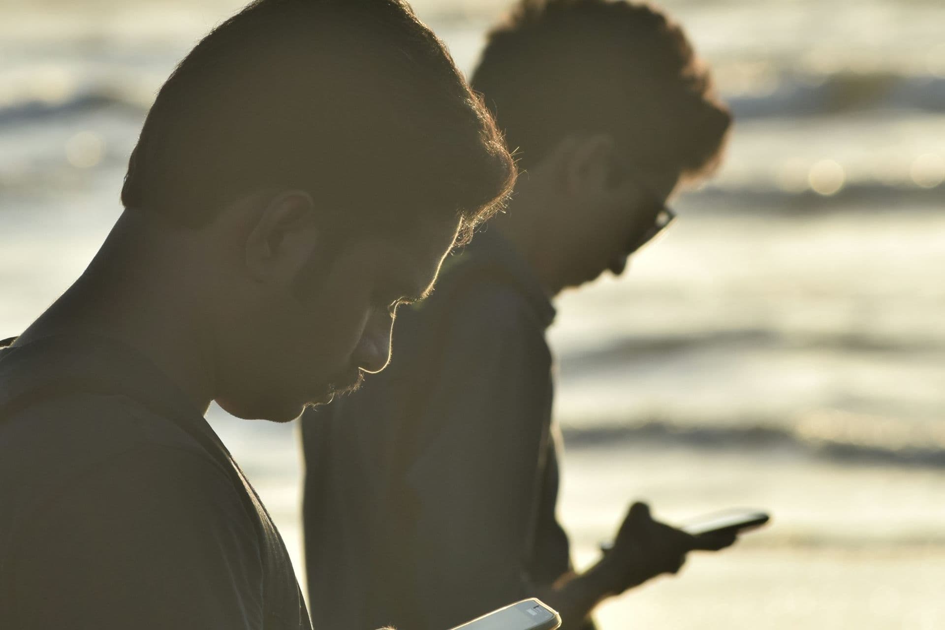 Two young men in silhouette looking at their smartphones on a sunlit beach