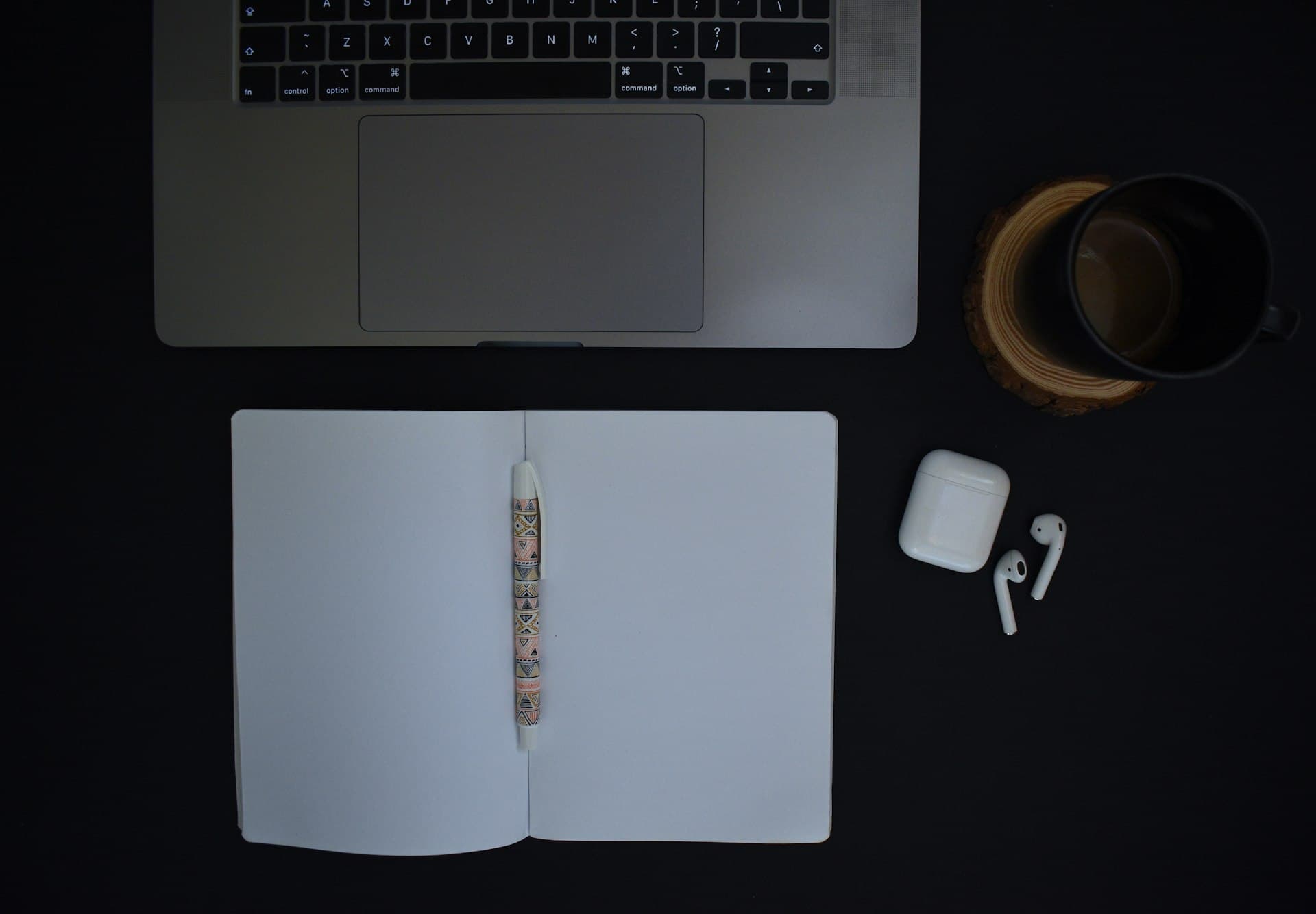 Top-down view of a laptop, open notebook, pen, coffee mug and earbuds on a black desk