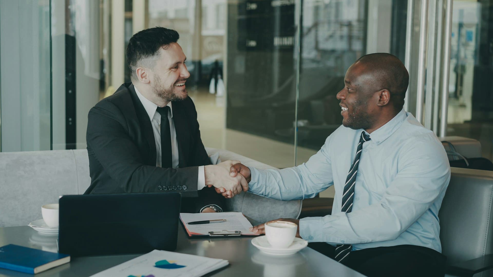 Two smiling businessmen shaking hands across a meeting table with coffee cups and documents