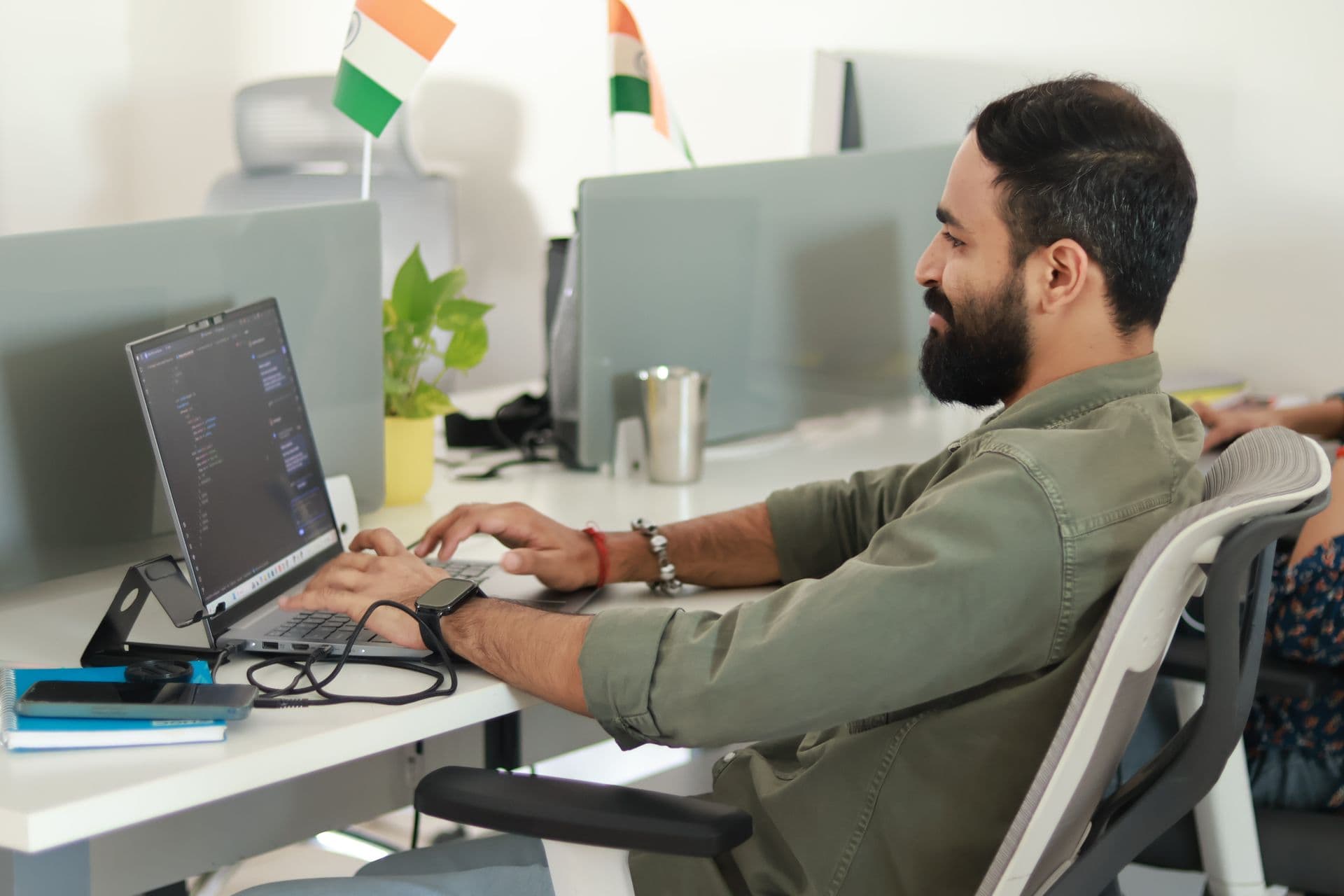 Developer typing code on a laptop at an open-plan office desk with small Indian flags