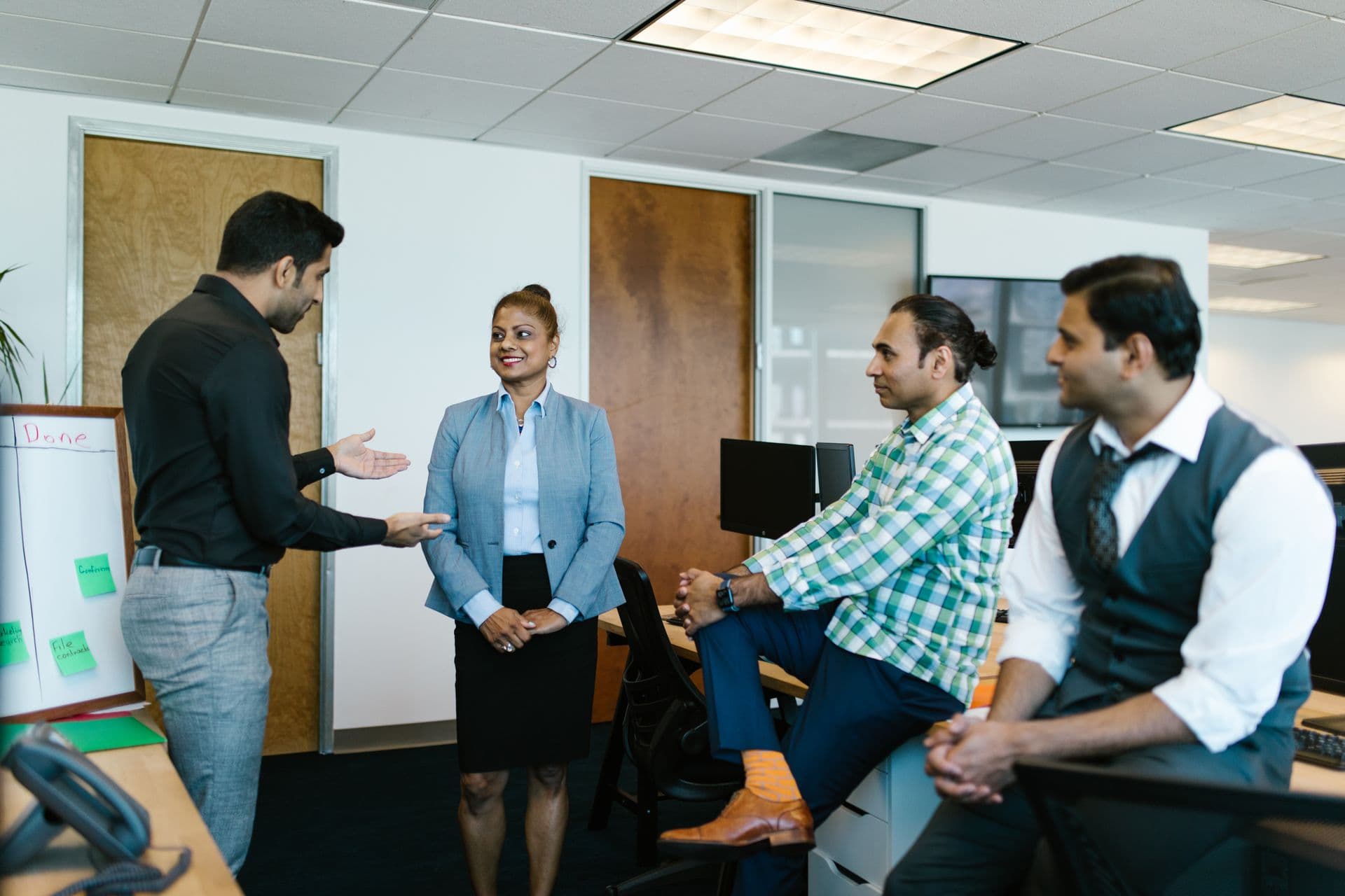 Colleagues in an open office listen as a man explains an idea beside a task board