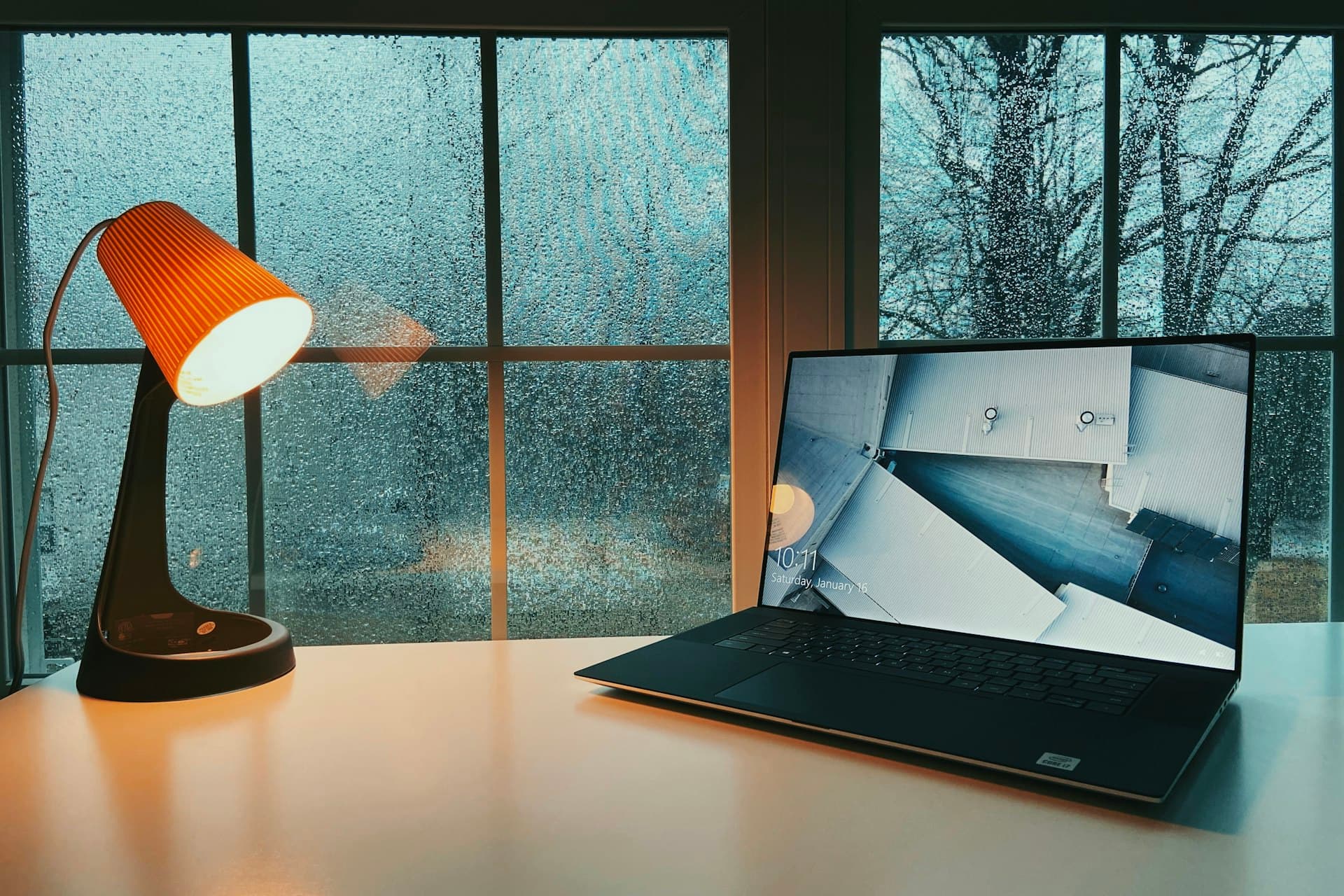 Laptop and glowing orange desk lamp on a table beside a window streaked with rain