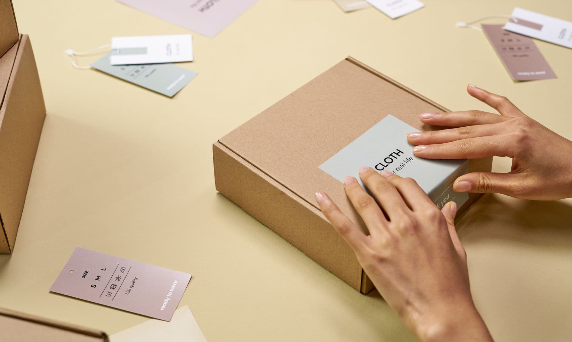 Hands pressing a label onto a kraft mailer box surrounded by swing tags