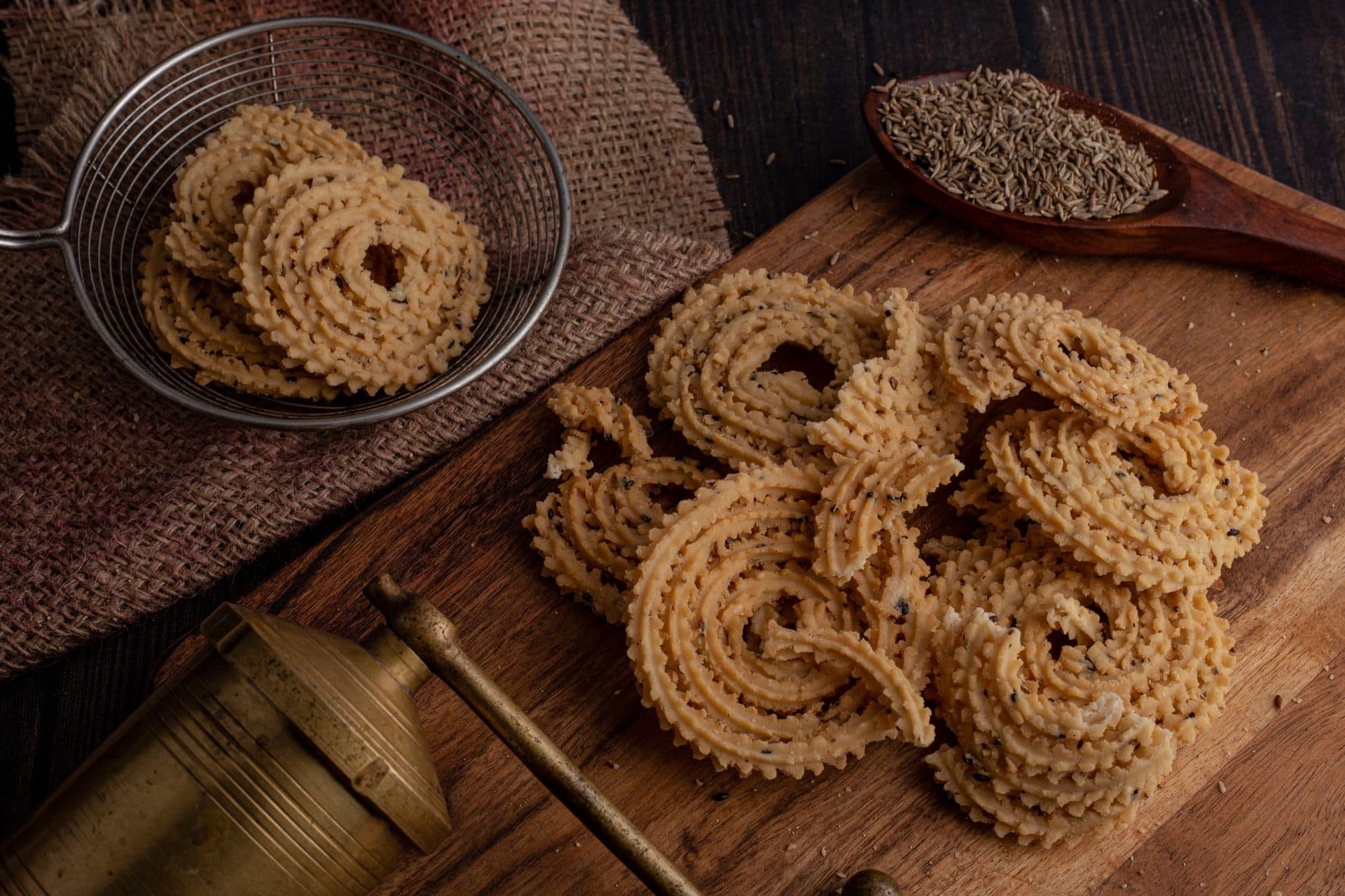 Golden murukku spirals on a wooden board with a sieve, cumin seeds and a brass vessel