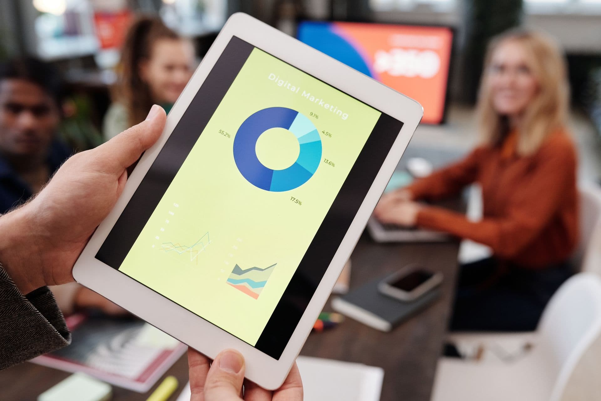 Hands holding a tablet showing a digital marketing donut chart during a team meeting
