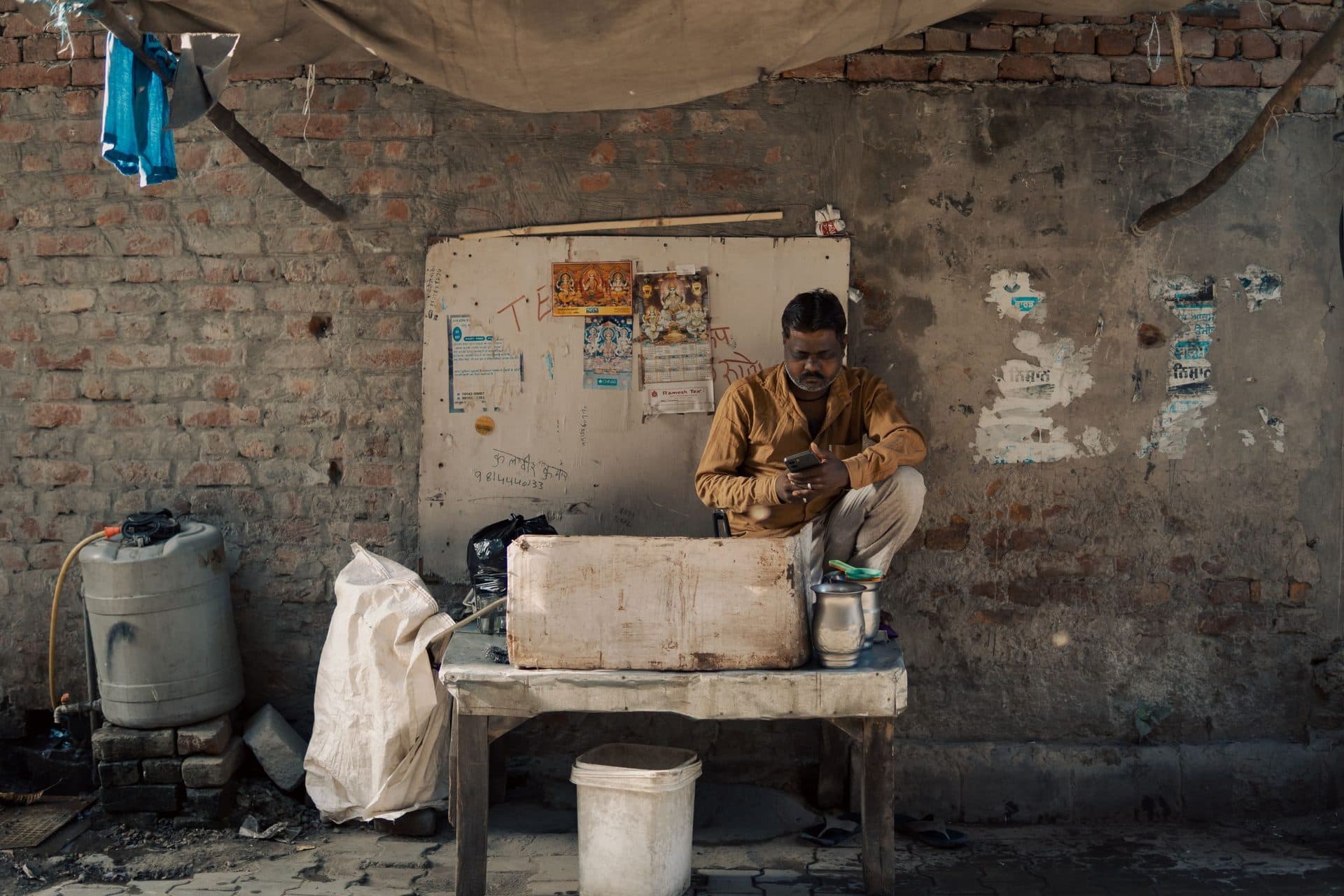 Street vendor seated at his stall checks his phone against a weathered brick wall