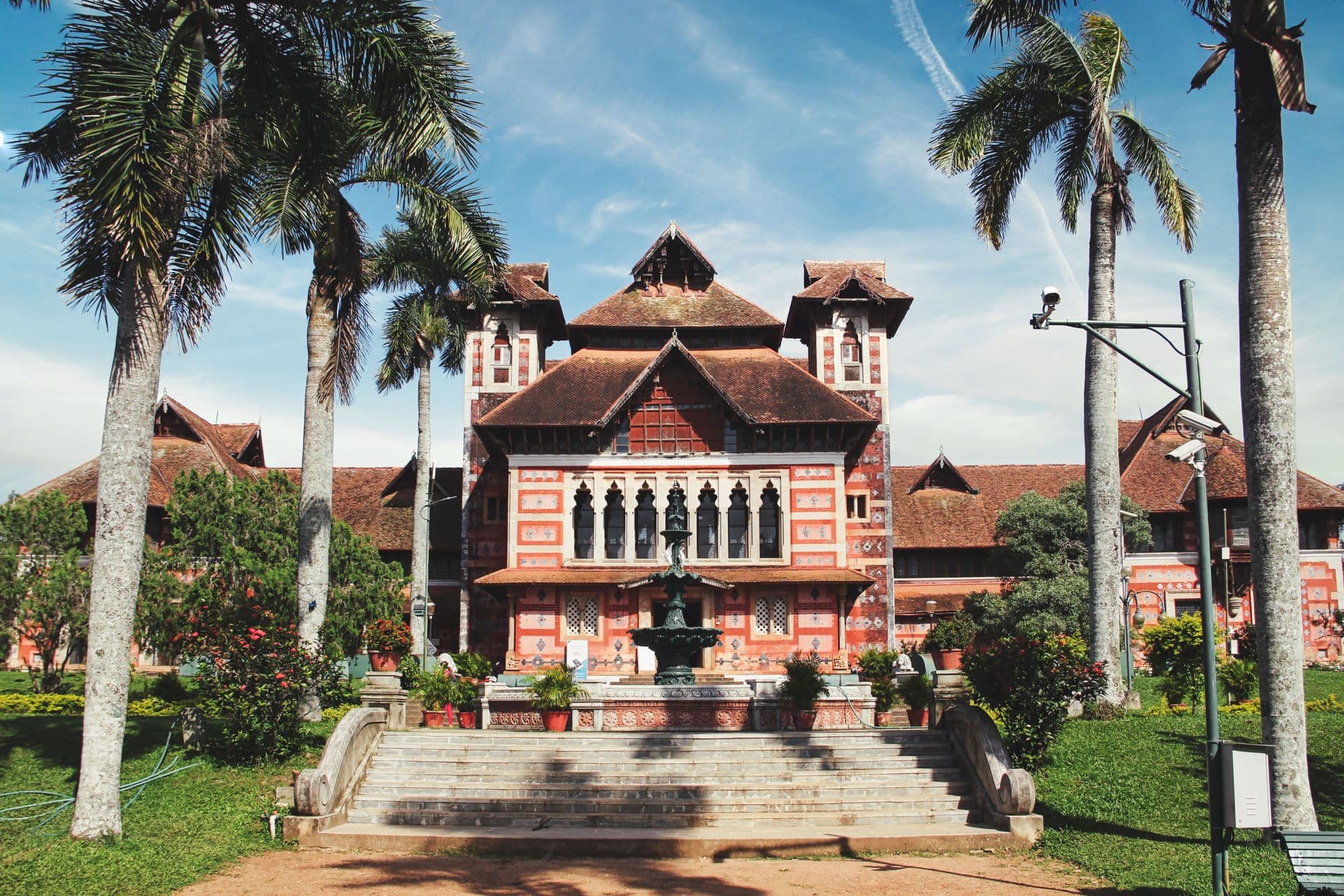 Napier Museum's red-patterned facade and sloping tiled roofs framed by tall palms and a fountain