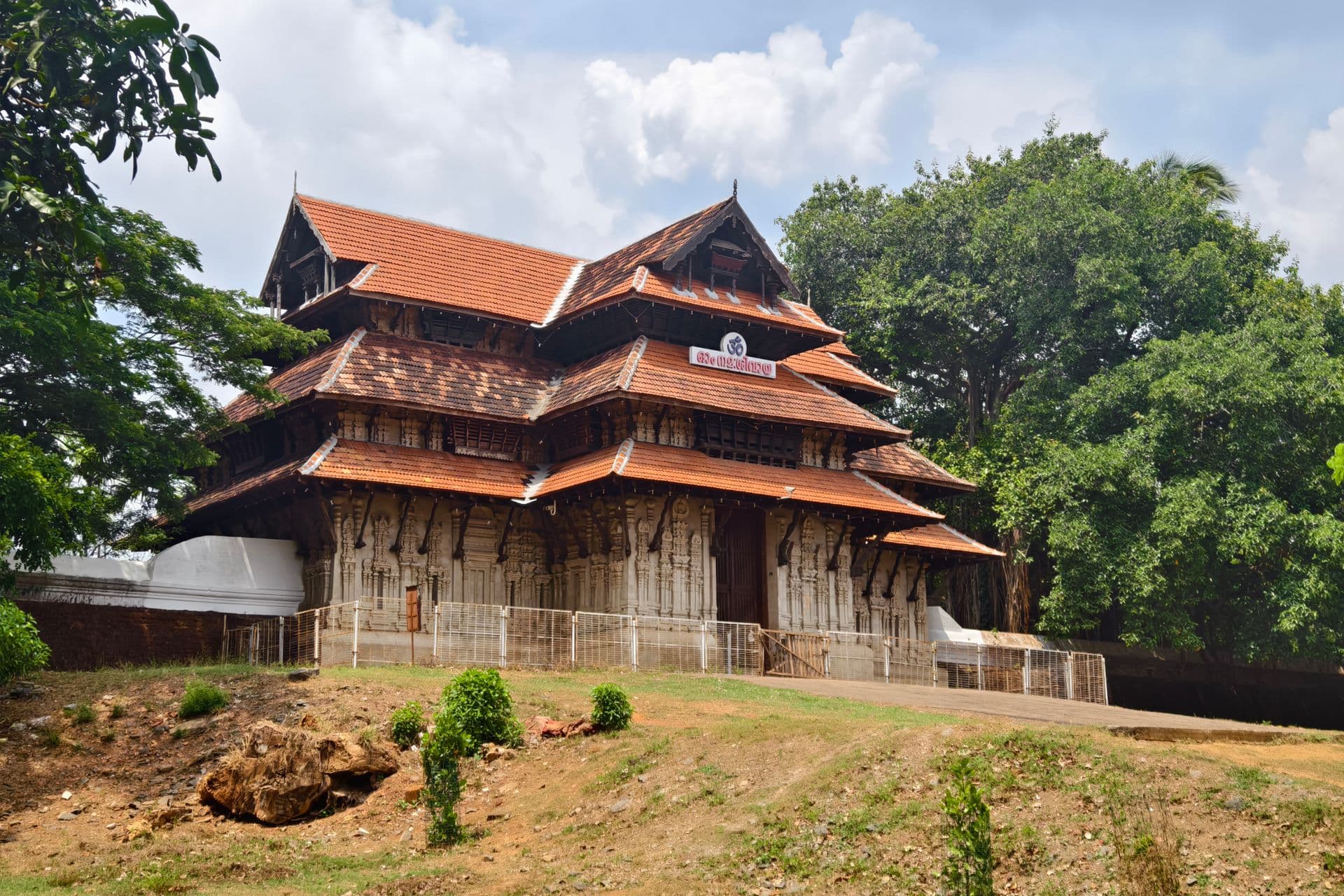 Tiered red-tiled roofs and carved walls of Vadakkunnathan Temple framed by large shade trees