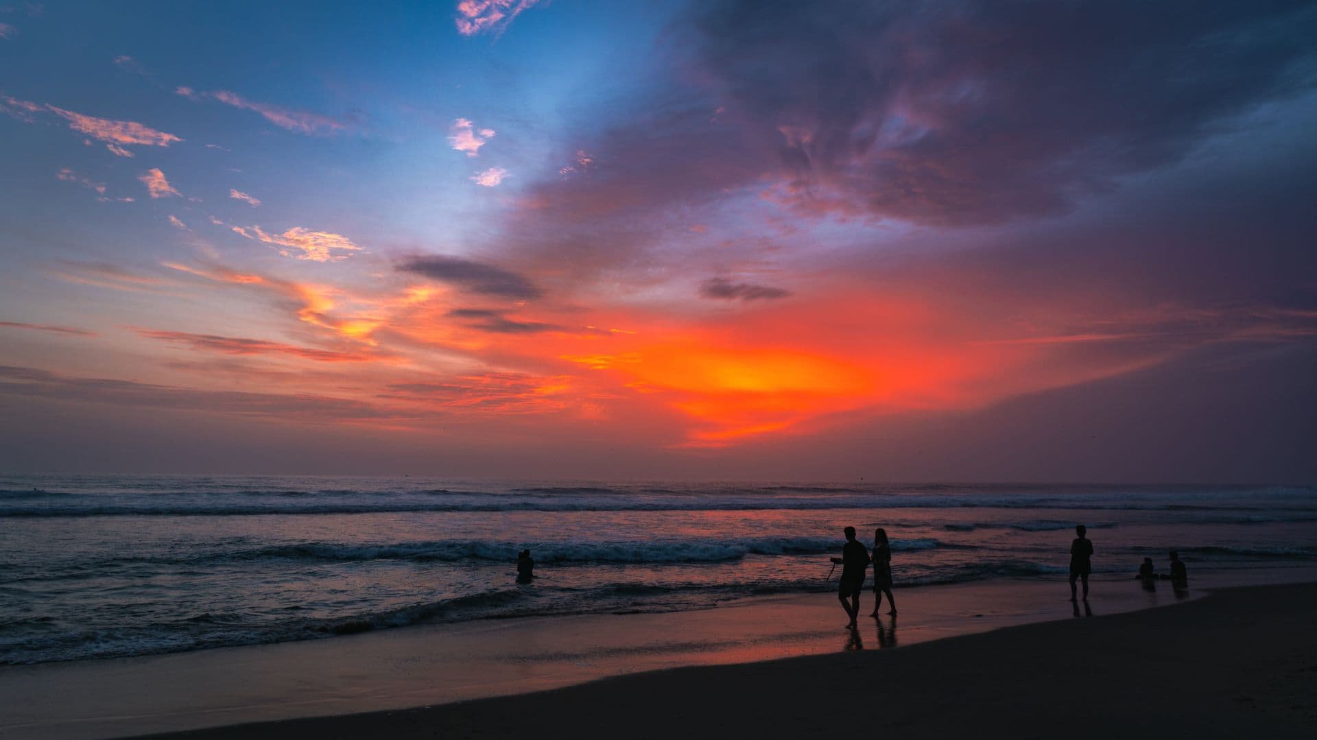 Silhouetted people on the shoreline beneath a vivid orange and violet sunset sky over the sea
