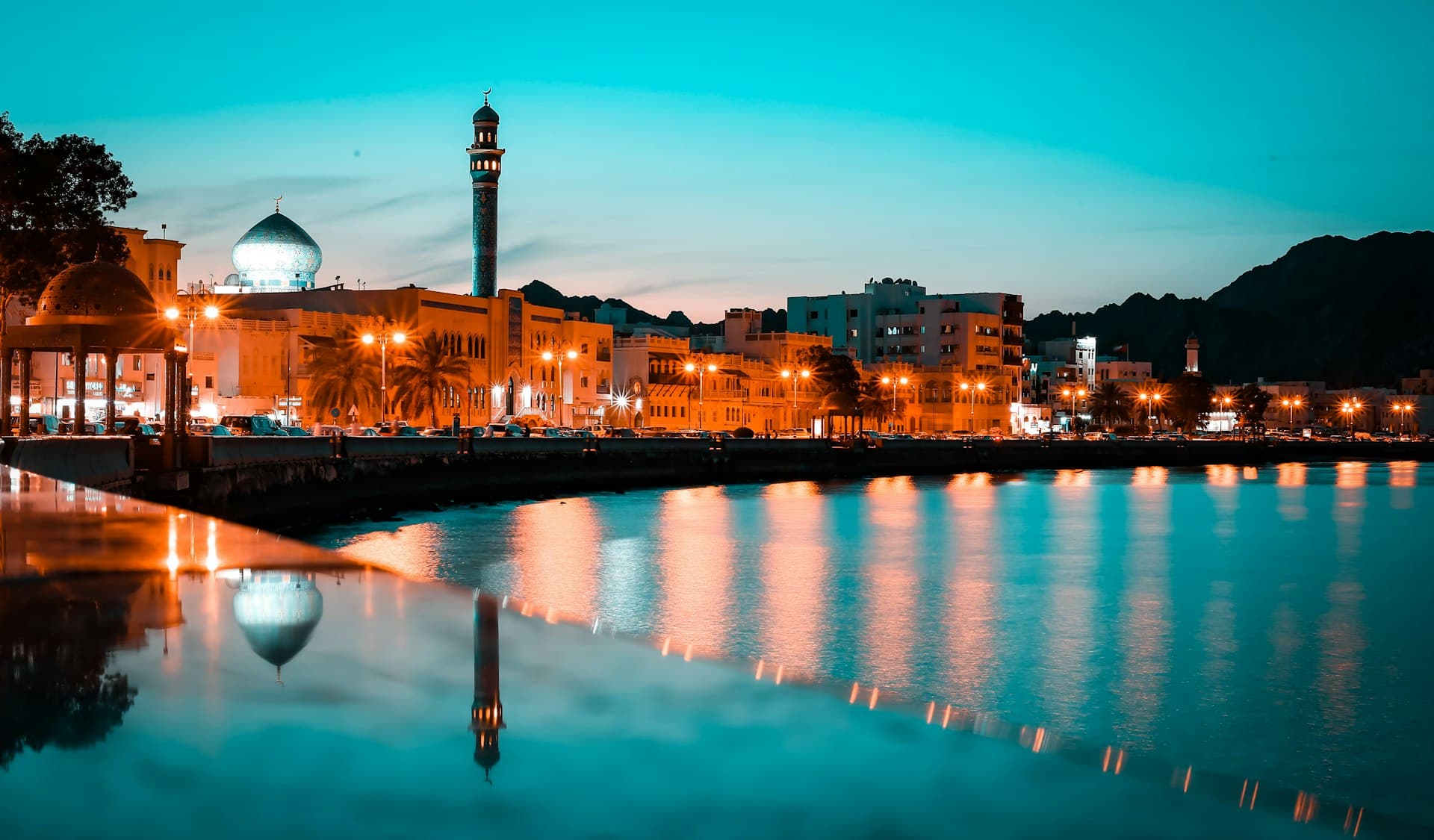Mutrah Corniche at dusk with a lit mosque dome and minaret reflected in water, Muscat, Oman