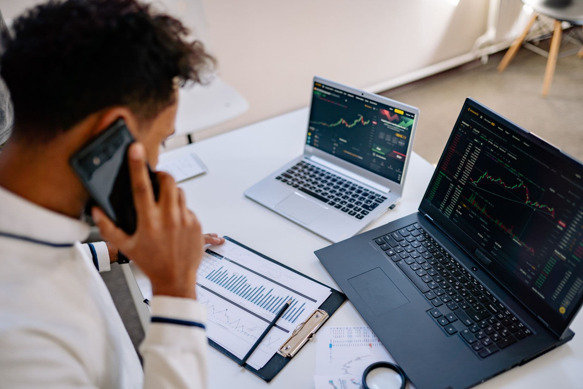 Man on a phone call at a desk with two laptops showing charts and printed reports