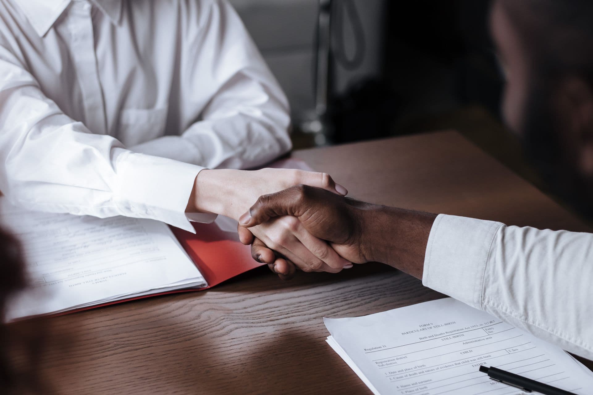 Two people shaking hands over a desk with documents and a red folder
