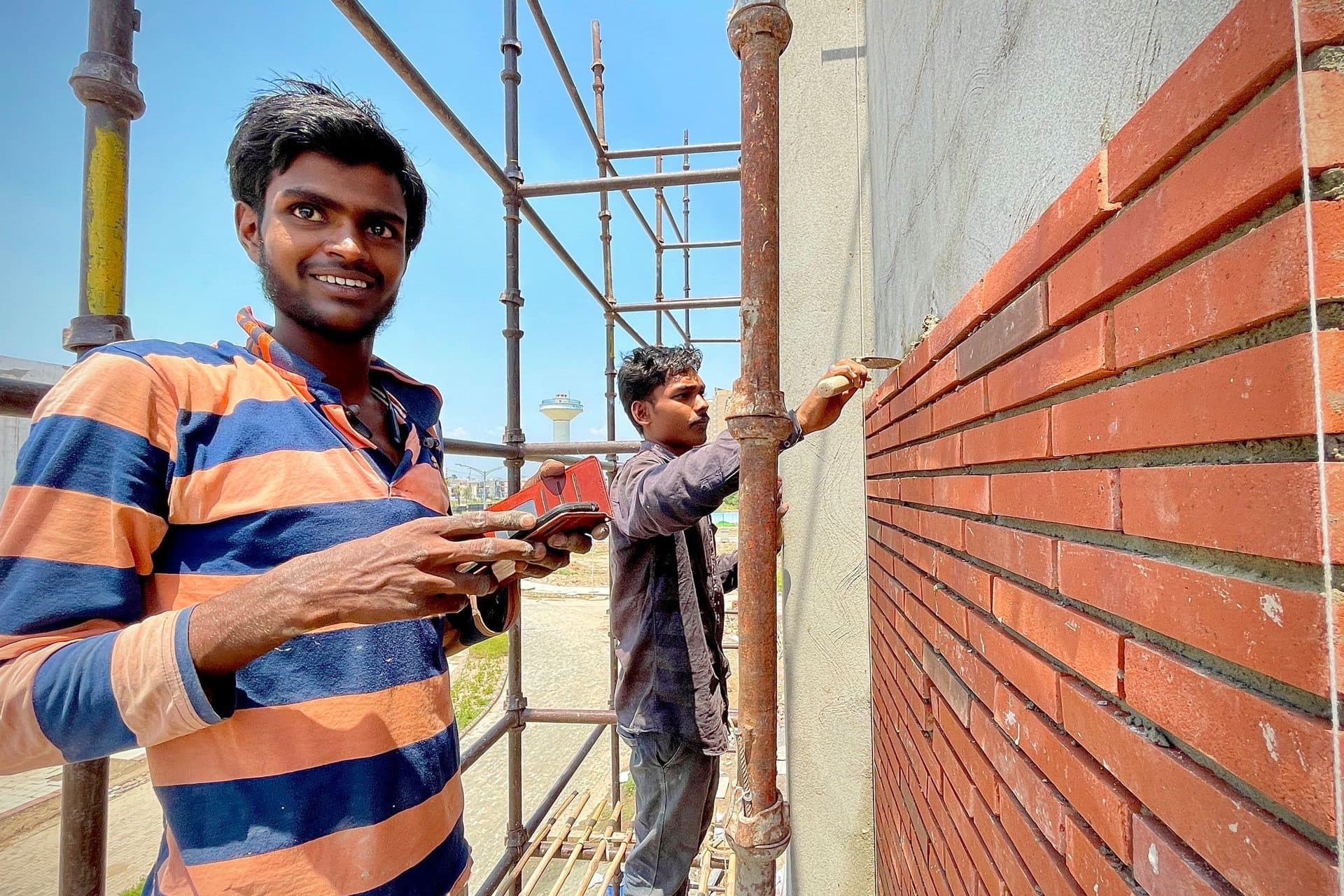 Two construction workers on scaffolding beside a red brick wall, one holding a phone