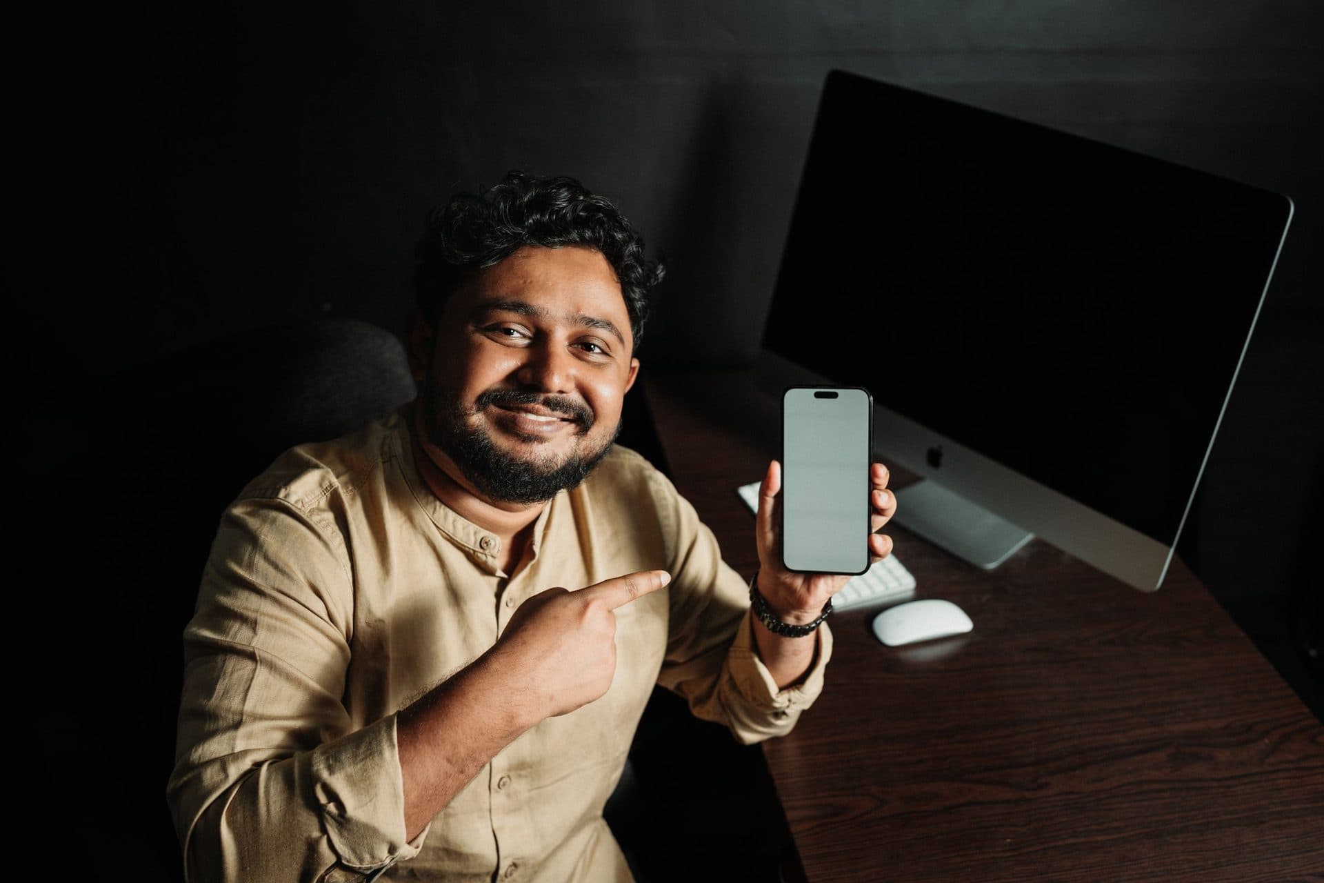 Smiling man points to the blank screen of a smartphone he holds up at a desk