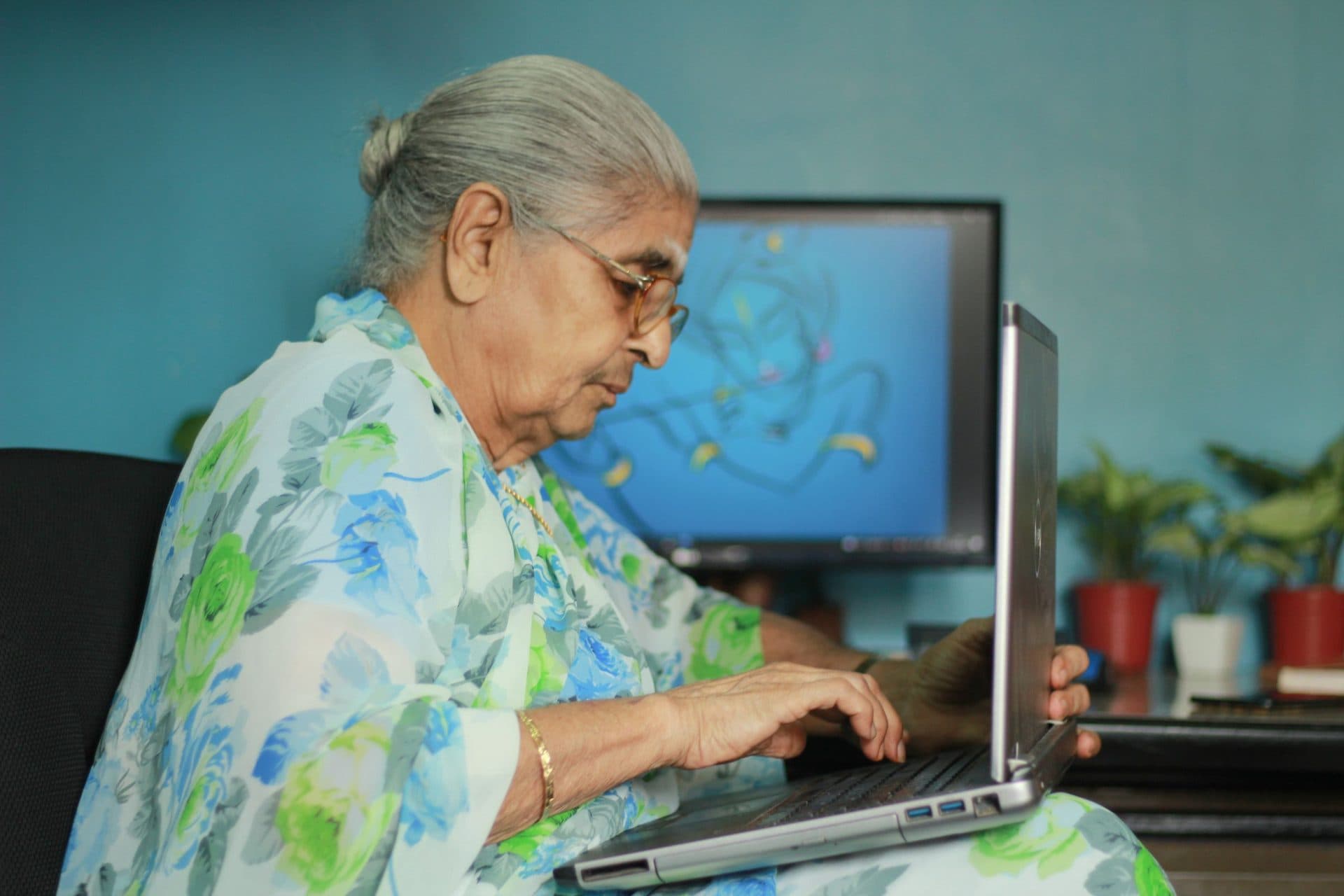 Elderly woman in a floral saree types on a laptop at a home desk