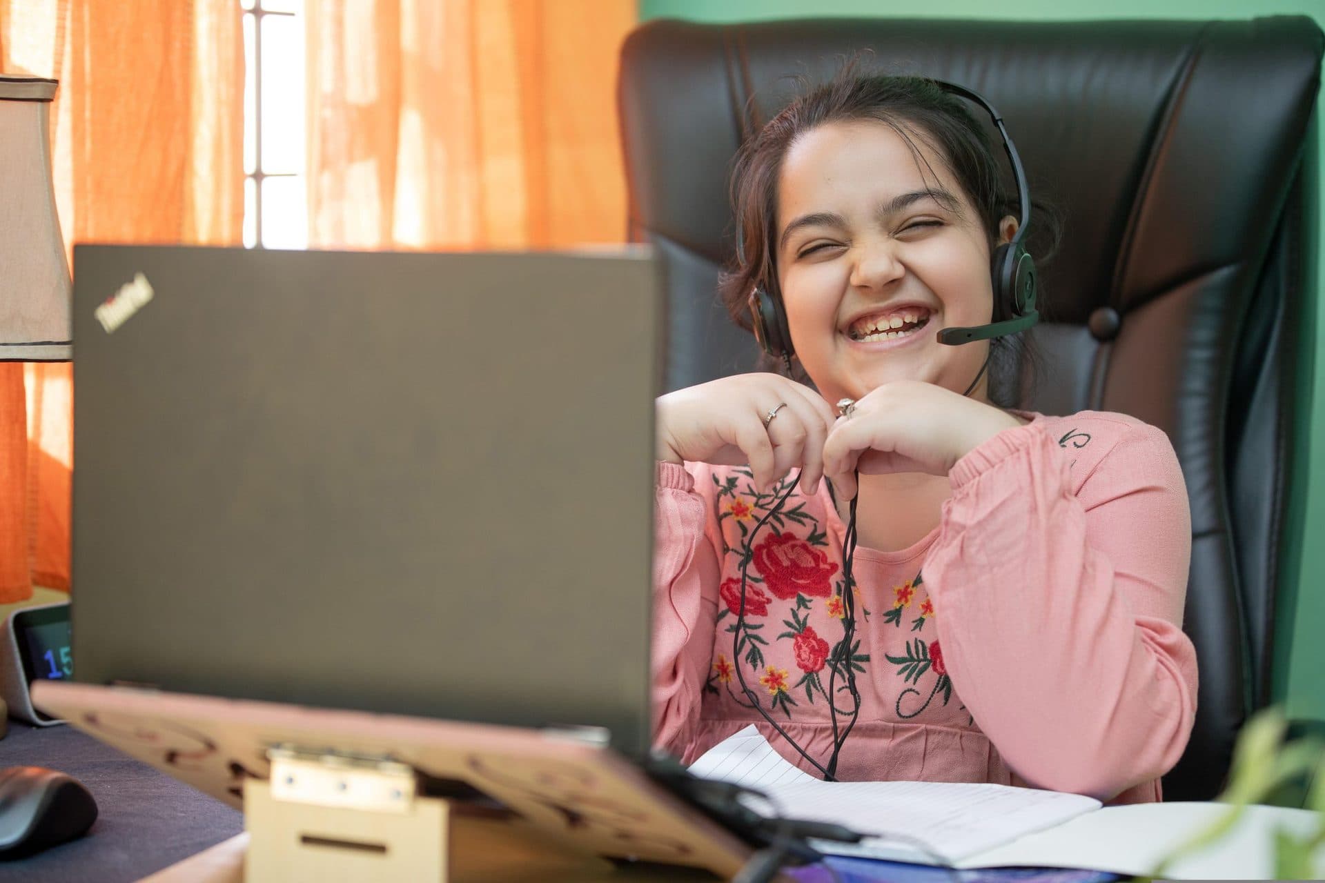 Laughing girl wearing a headset sits at a laptop in a home study area