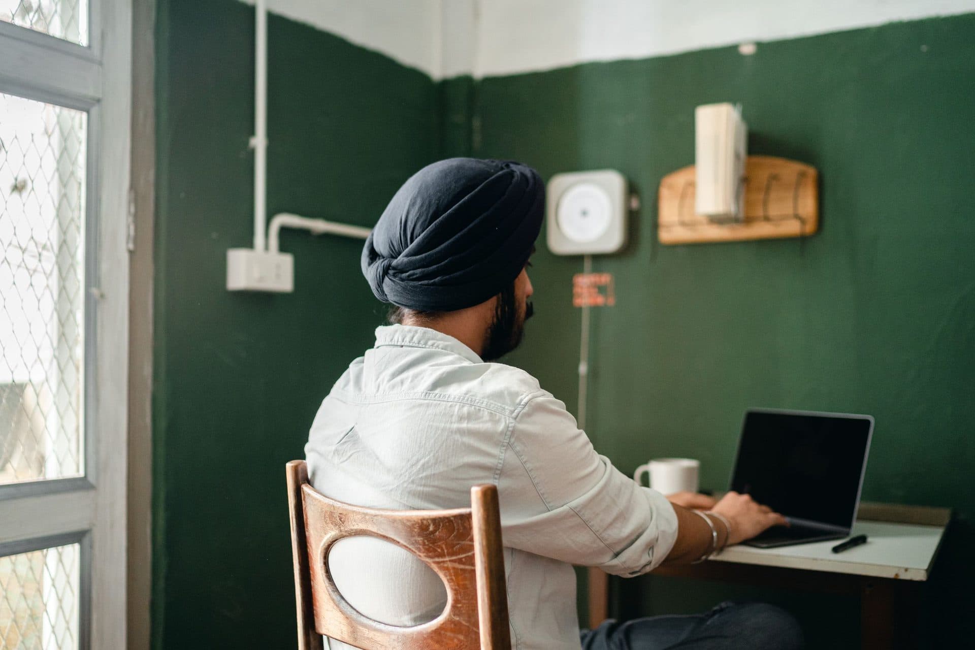 Man in a turban, seen from behind, types on a laptop at a small home desk