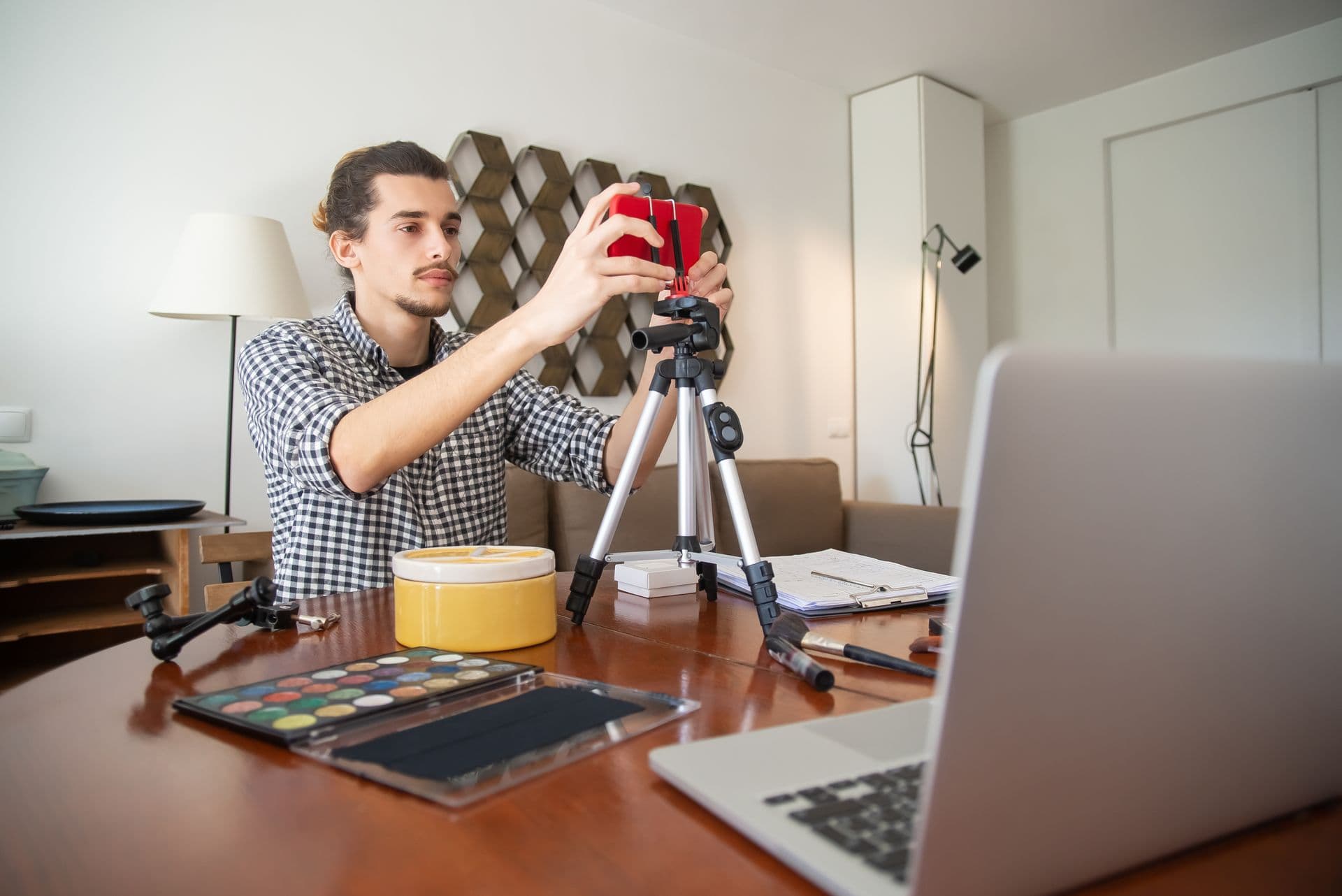 Man mounting a red smartphone on a tripod at a desk beside a laptop