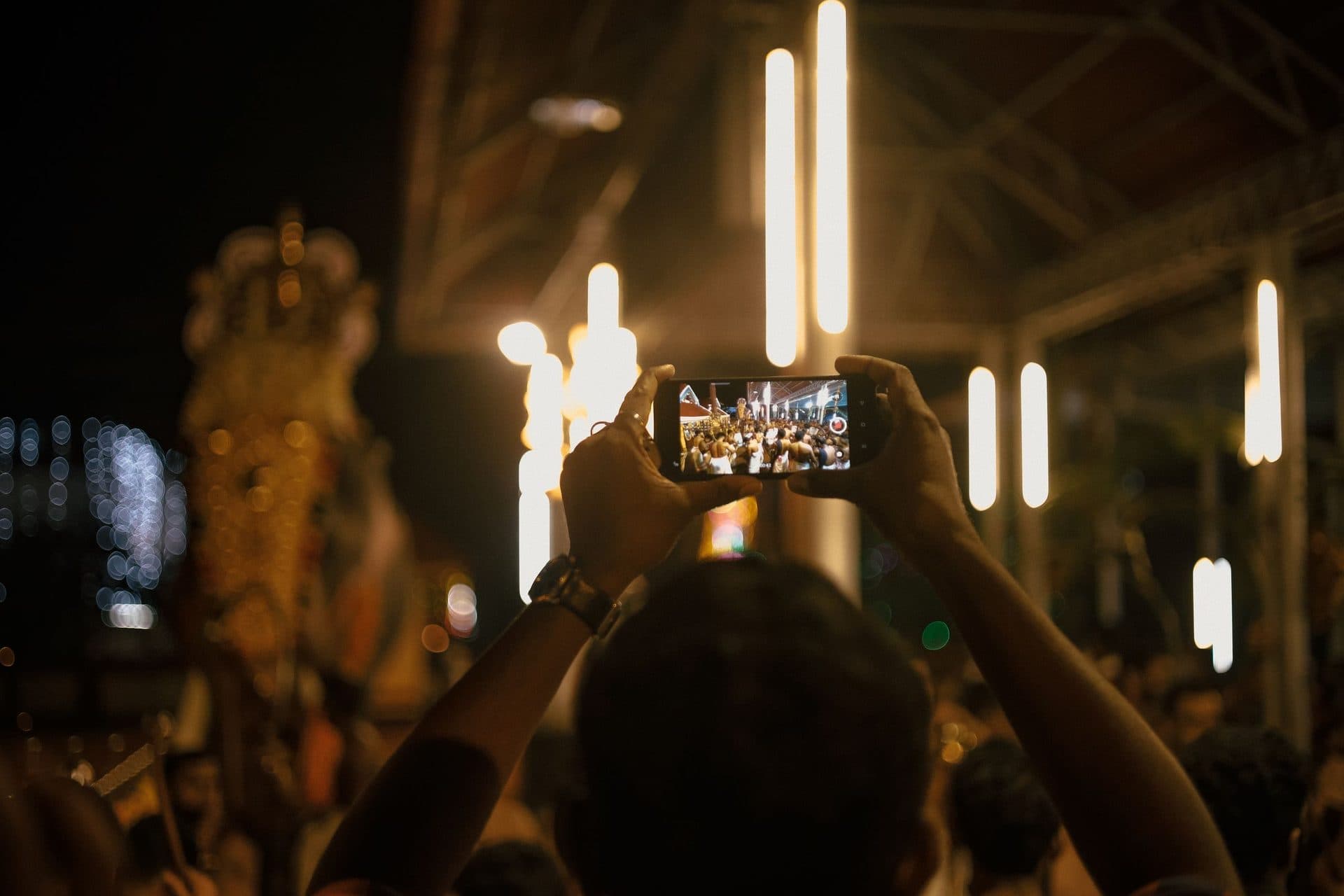 Raised hands hold a smartphone filming a crowded festival at night under bright lights