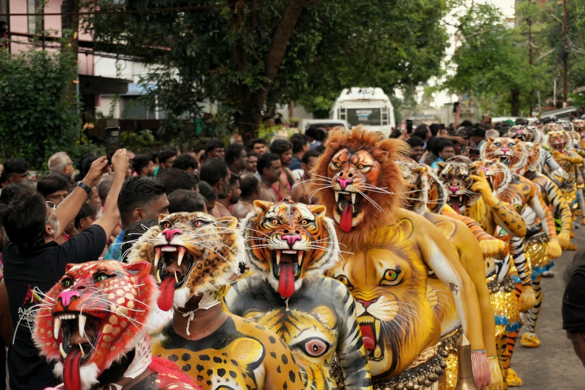 Tiger-painted Pulikali performers parade past a crowd, some spectators filming on phones
