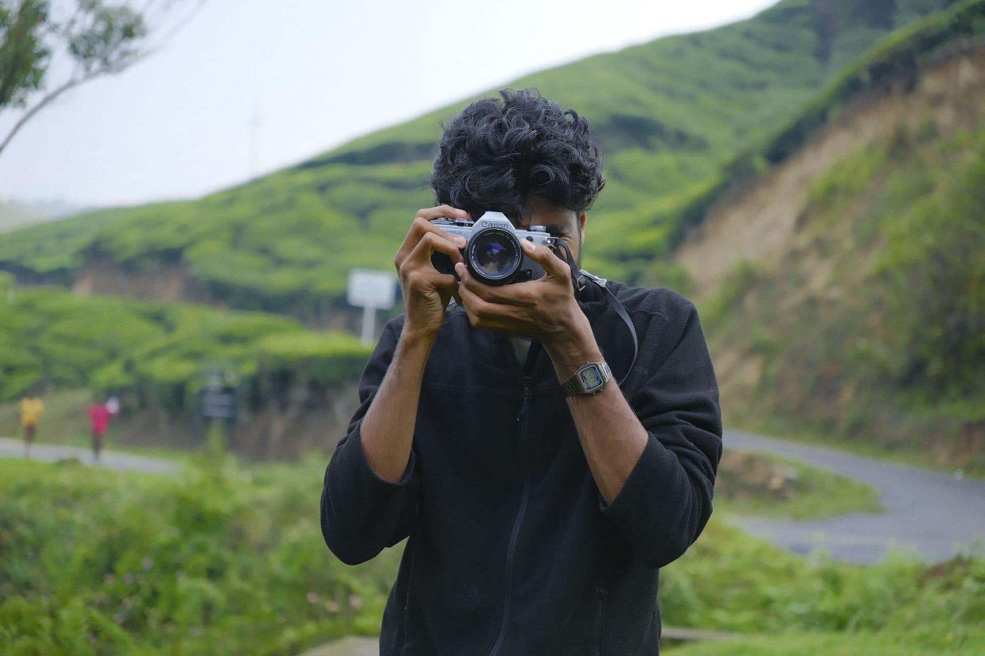 Young man aims a silver camera at the viewer on a road through tea-covered hills
