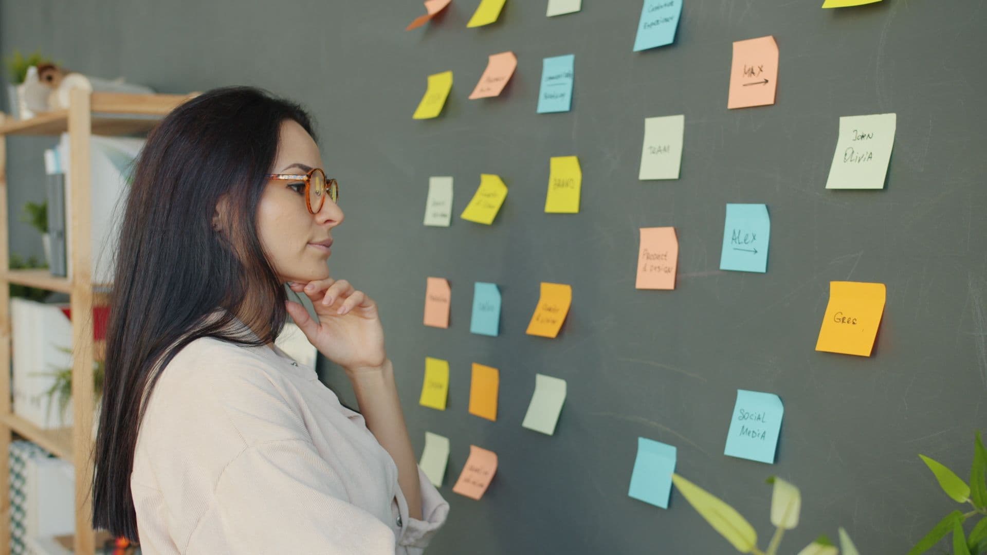 Woman in glasses studying colourful sticky notes arranged on a chalkboard wall